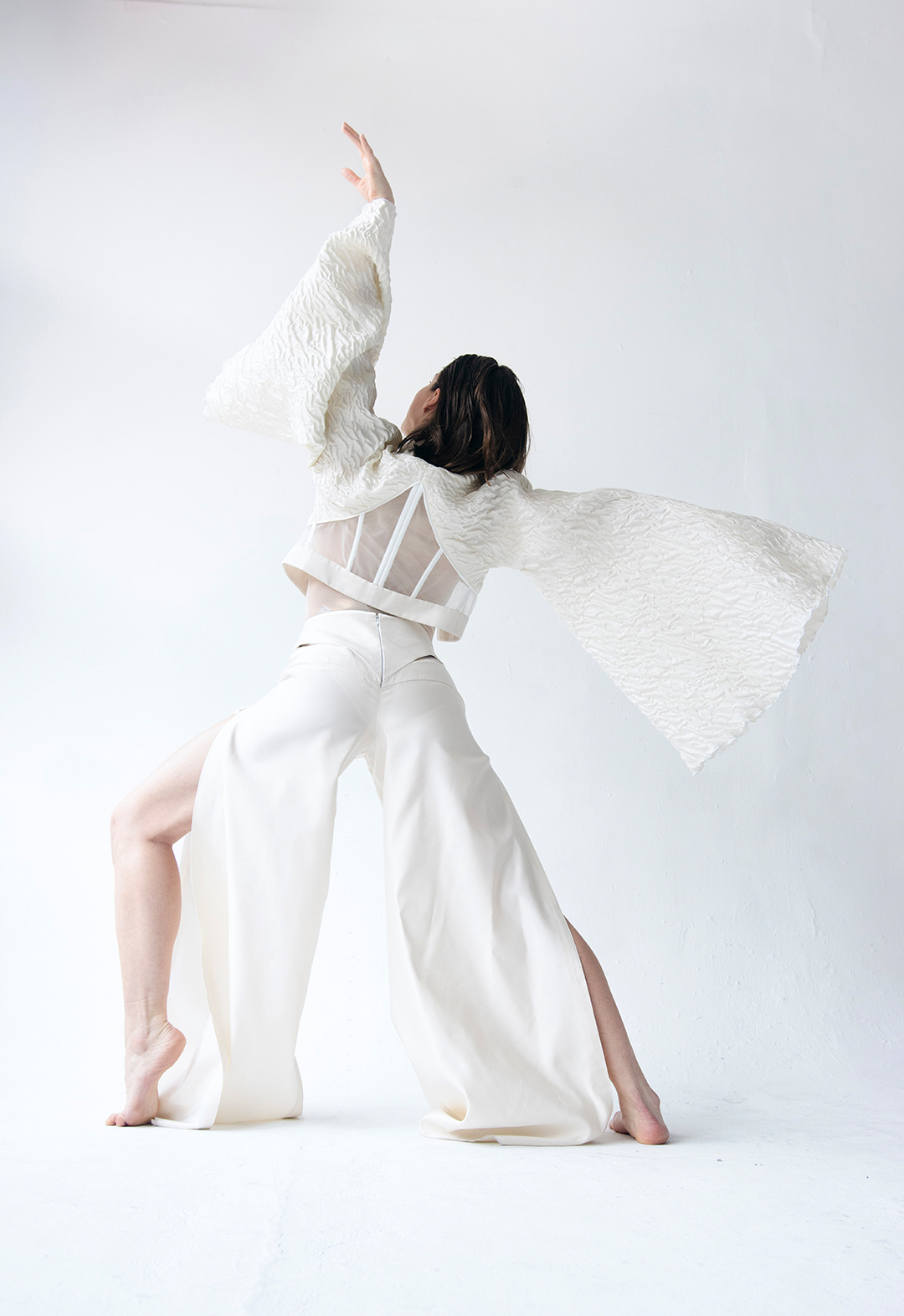 Back view of a model wearing a seashell white corseted jacket with flowy kimono sleeves and flared, cut-out, and slitted pants. Her left hand is up in the air and the right hand is sticking out towards the right, parallel to the floor. Her left knee is bent and the right leg stretches out diagonally back. 