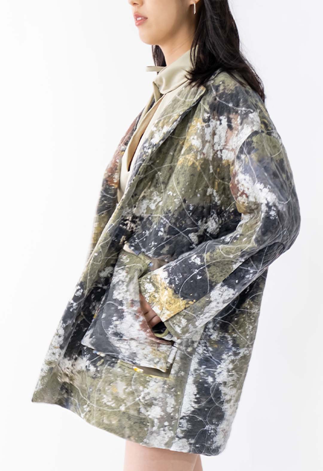 Side view of an abstract scrap fabric jacket, showing the details of a cut-out pocket that is held together with tabs on both the jacket and the pocket itself. Two tabs are held together on each side of the pocket with hardware made from resin that has flowers pressed inside. The model is in front of a white background and is looking off into the distance with her hand inside the pocket.