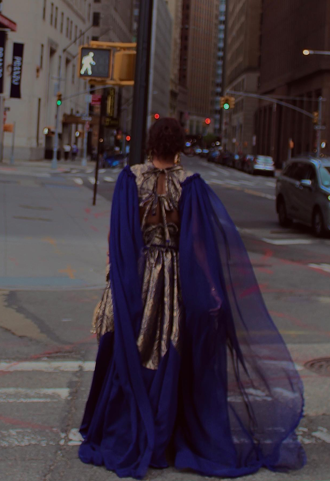 Back view of a model walking on the streets of New York City. As the model moves, the chiffon sleeves gracefully flow behind, drawing from the inspiration of royal capes. You can also see the tie details of the open back vest which is completely different from the front. 