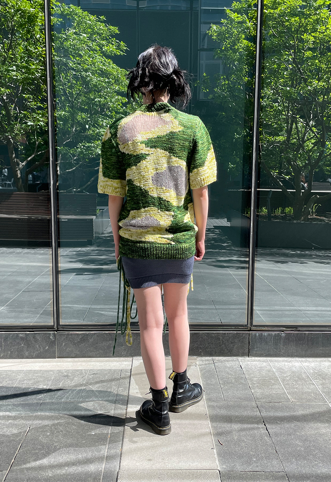 Back view of a model wearing a hand knit short sleeved intarsia sweater. There is a glass building in the background with reflection of trees.