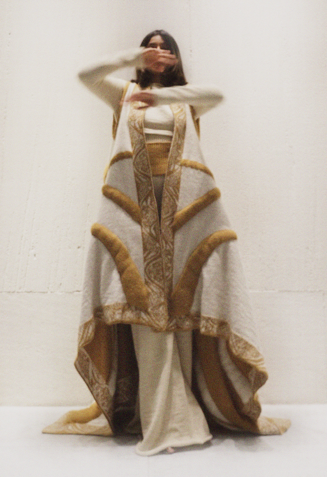The model is wearing a huge two-color, jacquard pattern reversible cardigan, which has borders all around. The tan pattern lines inspired by sari are stuffed. She is wearing white wide-legged pants, which are a little bit visible below the hemline of the cape. Her hands are up in the air, and the background is white.