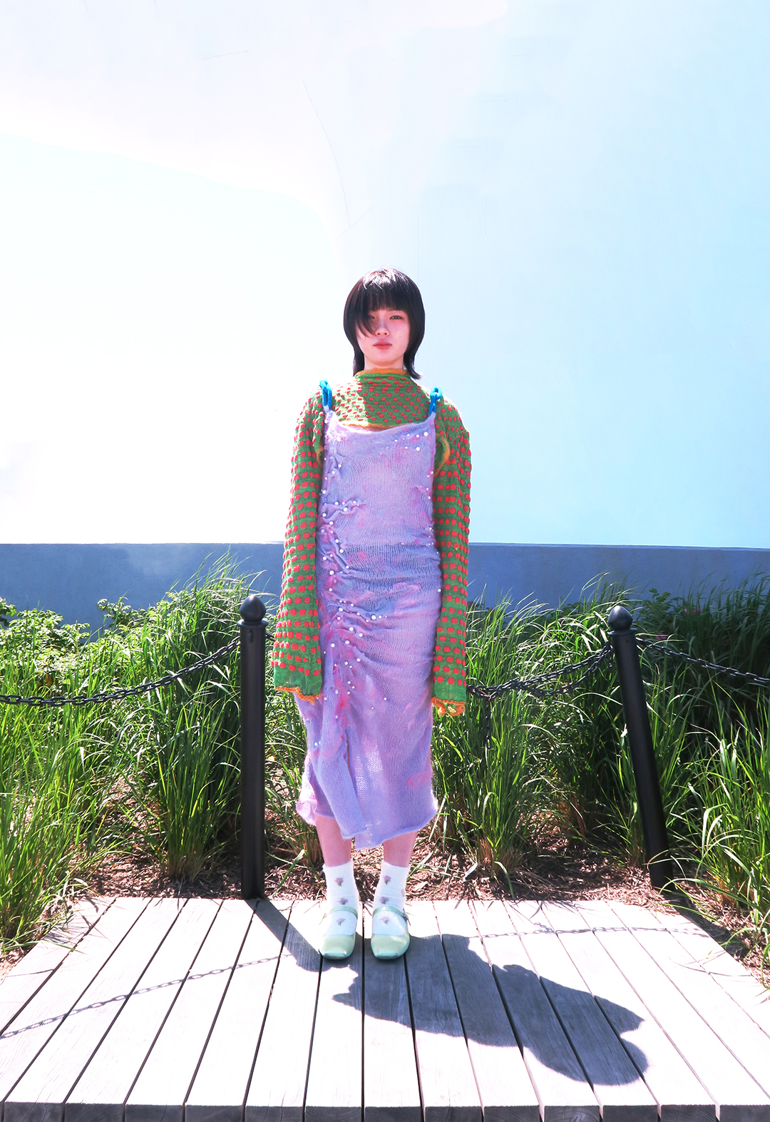 Front view of a model wearing a green sweater with red polka dots and a pink spaghetti straps knit dress over the sweater. The model is standing on a deck and there is tall grass in the background.