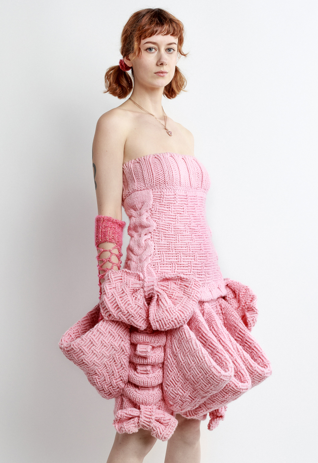 Photo of a girl posing in a pink sleeveless magical girl mini dress, looking to the camera. The dress is hand knit with baby pink wool. The dress features big bows and big pockets circling the skirt of the dress. The dress, and girl, are posed at a three-quarter view to the camera. The girl is wearing crochet fishnet gloves, pigtails, and a heart necklace. The dress is angled so that the biggest bow trim is directly in view.