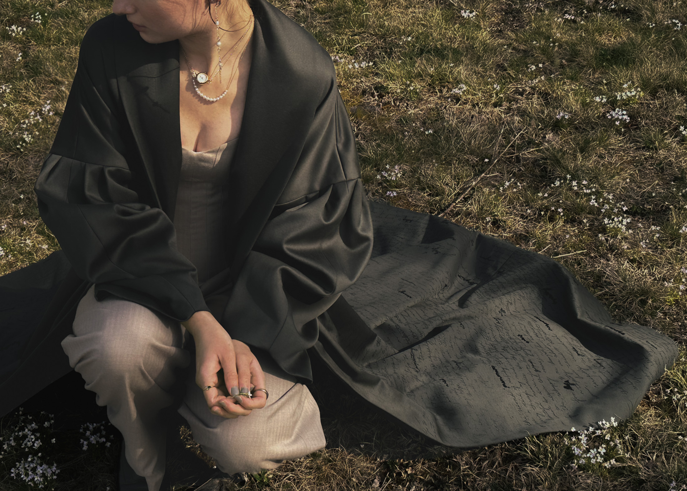 Photo of a girl wearing a green coat and beige jumpsuit and kneeling in the grass with small white flowers around her. The hem of the coat is spread out behind her and contains the imagery of words.