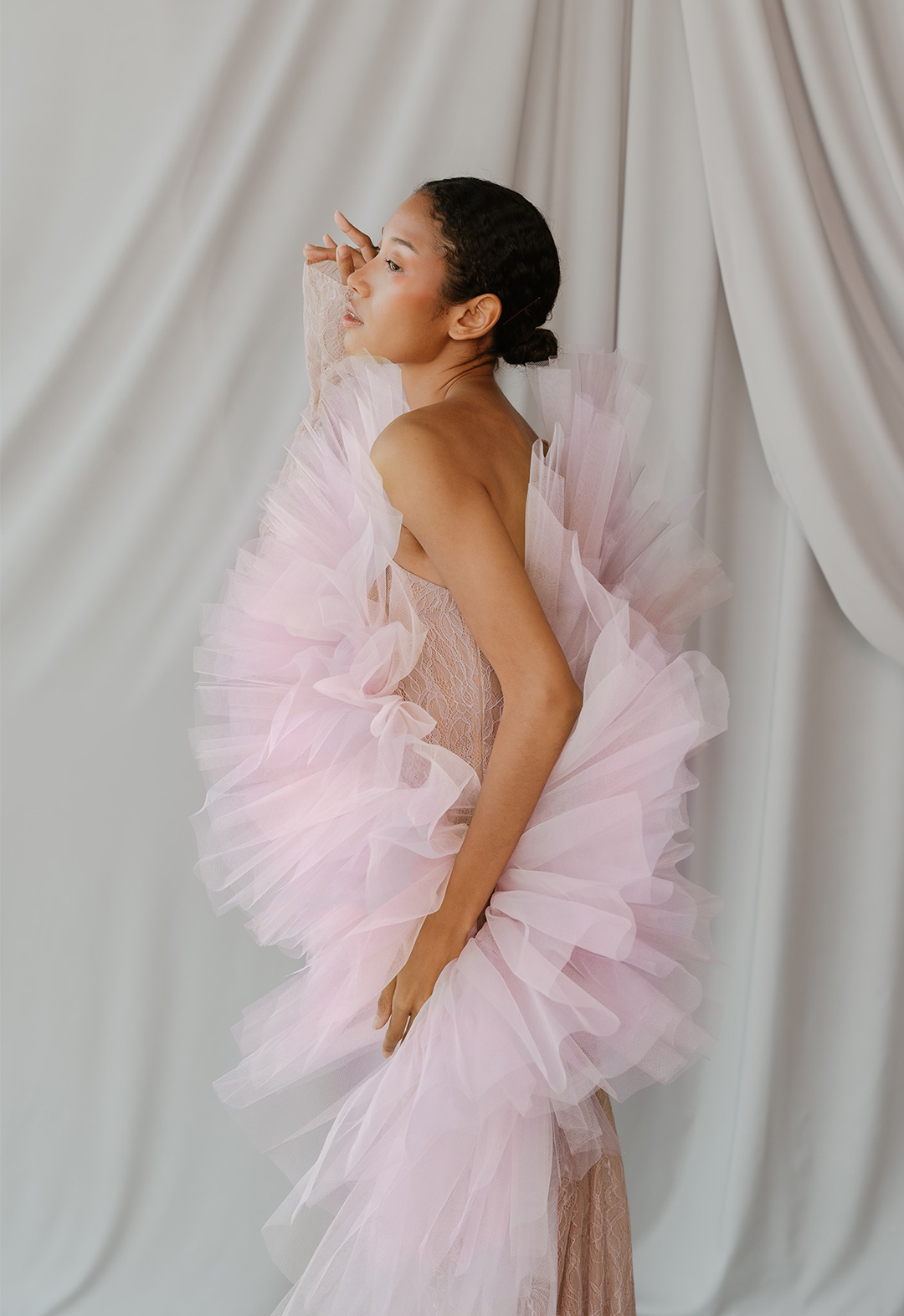 Side view of model in blush lace, one-shoulder sleeve dress with godet train, lace appliqué, and pleated tulle coming from a curved seam over the bust.