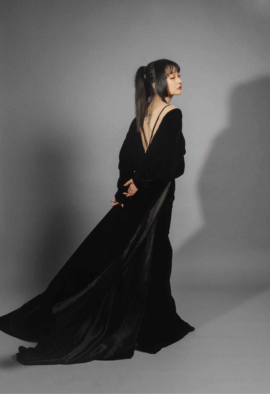 This black velvet dress has a V-shaped open back and narrow straps on each shoulder for support. The dress is long, with a train that flows behind the wearer. The background in the image is gray.