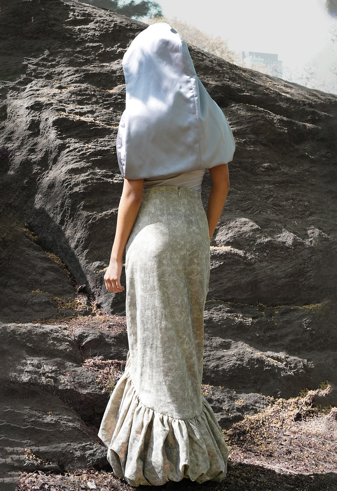 Back view of a model wearing a curved bubble skirt and a hooded capelet. There is a big rock in the background.