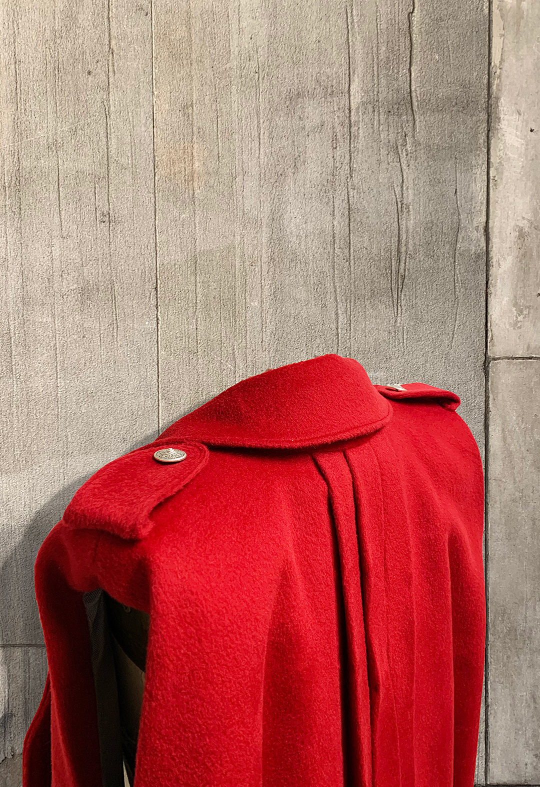 This is the back of a red wool vest. There are two metal buttons on top of the stripes on the shoulder. The background is a concrete wall.
