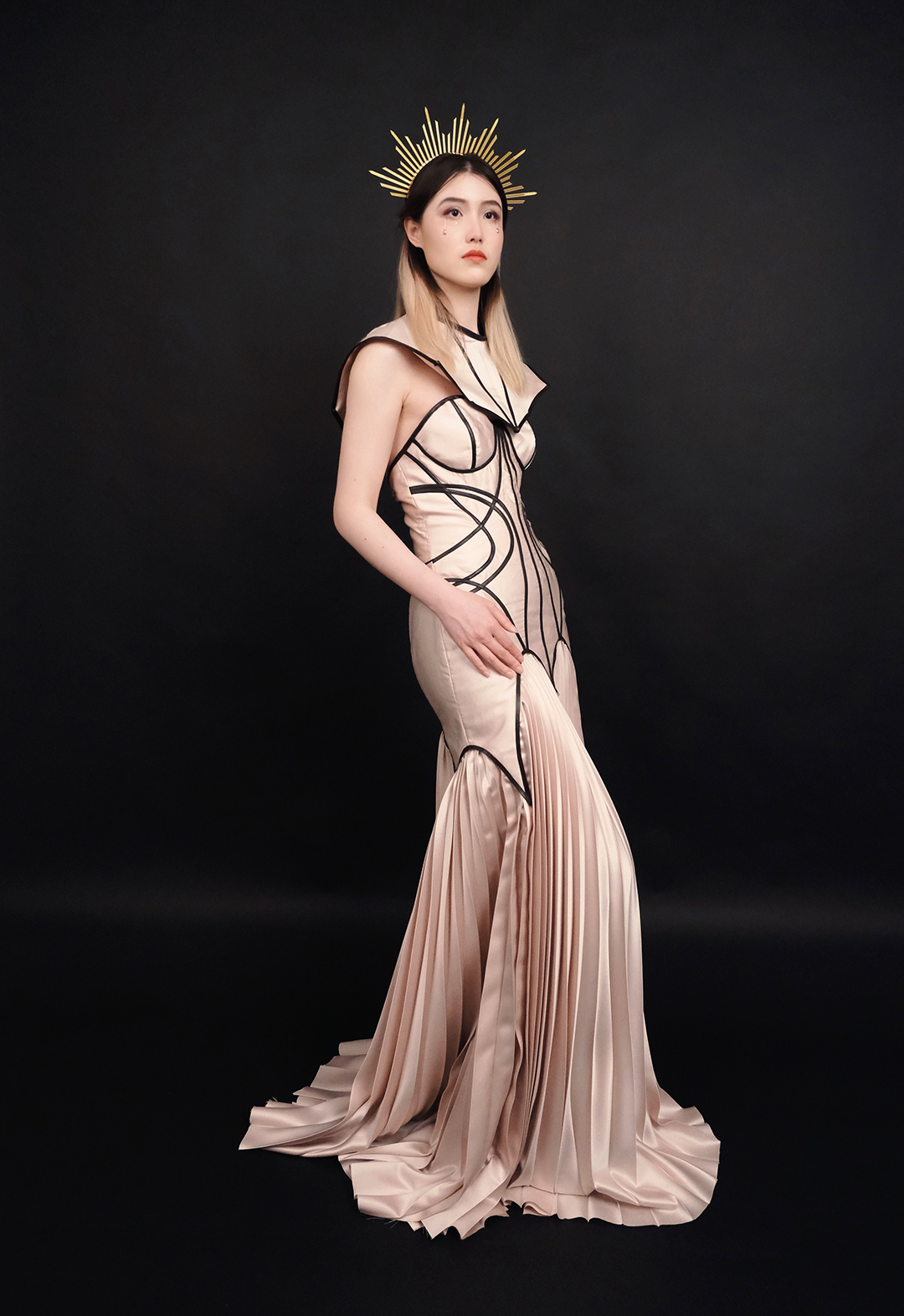 Side view of a model wearing a mermaid gown with fitted torso and pleated godets in the bottom. The model is wearing a hairband that gives a halo look. She has her visible hand on her thigh.