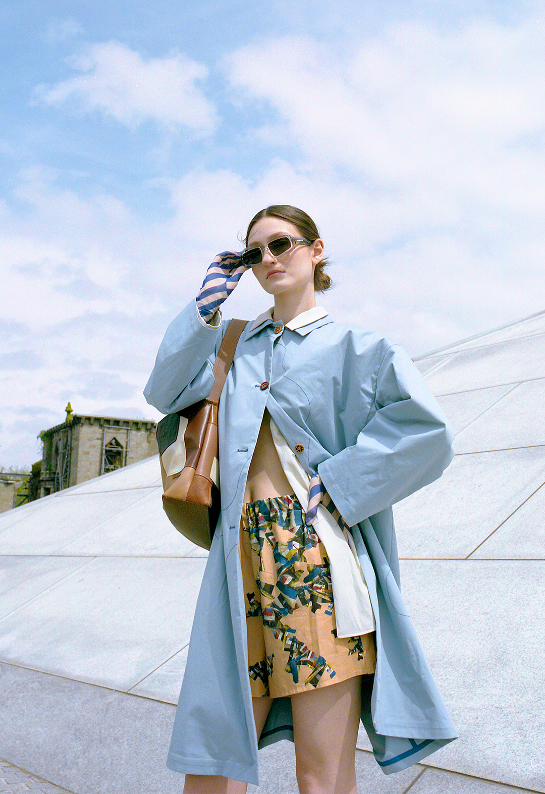This is a long, blue oversized coat, with flower-inspired embroidery detail and ethnic wooden vintage buttons. Worn underneath with a green, embroidered oversized shirt and patchwork digital printed shorts. Accessorized with dark-blue mesh, long-sleeved gloves, sunglasses, and a handmade patchwork tote bag made out of recycled scrap leather. The model is posed with her hands above her head in front of a gray wall.