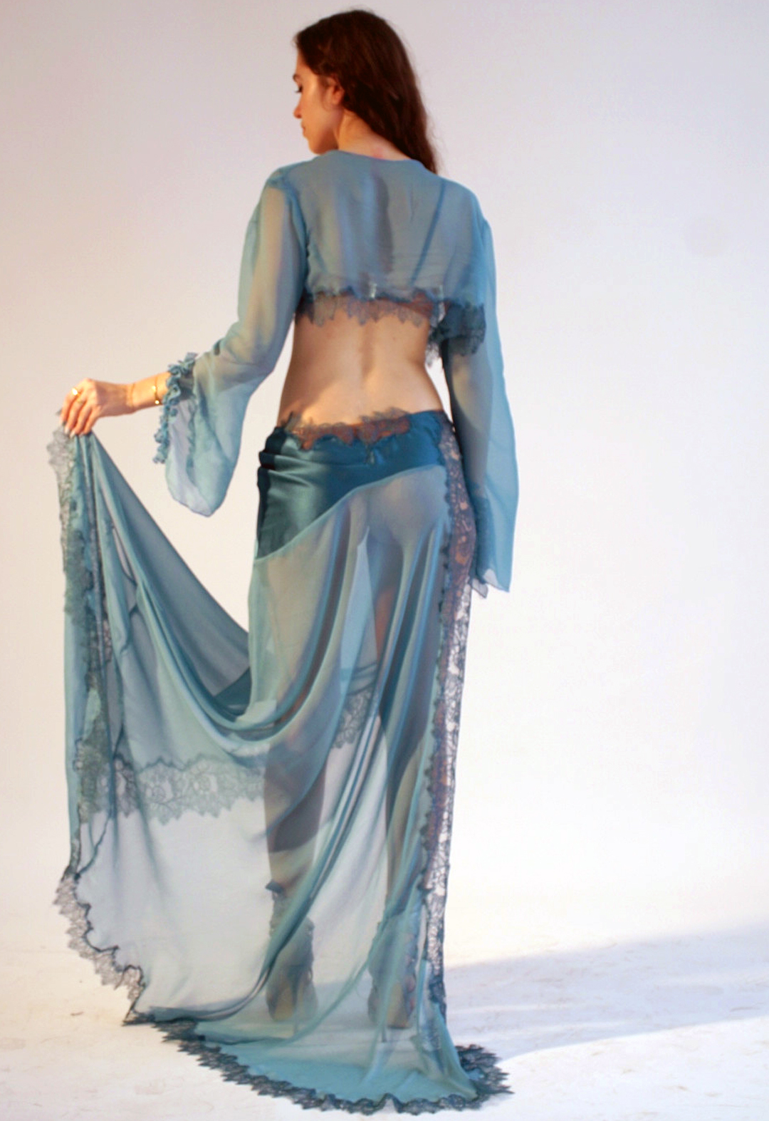 The model is facing away from the camera with her hair down. She is holding the slit of the skirt, giving a draped ripple effect to the flowing chiffon. Her hair is pushed to the right and she is looking down toward her hand. Copper double-layered mesh soft bralette with lace appliqué at the side seam. Gold hardware is used on the clasp on the back, the rings, and the sliders. Deep teal–colored silk adjustable straps hold up the bandeau-like, rounded shape bra. Deep-teal silk skirt. Crepe silk charmeuse asymmetrical yoke, resembling a wave, reveals a built-in cheeky panty within the skirt. The skirt is continued by a long flowy drape of the same colored silk in chiffon. The skirt has a slit and is hemmed with hand-dyed lace, cut asymmetrically, to resemble abstract oceanic features. There is a cropped chiffon bed jacket, as a two-in-one cover-up. The jacket has a slit at each sleeve, and the slit has a purled edge ruffle at each side of the slit. Hemmed with hand-dyed lace cut asymmetrically, to resemble abstract oceanic features. 