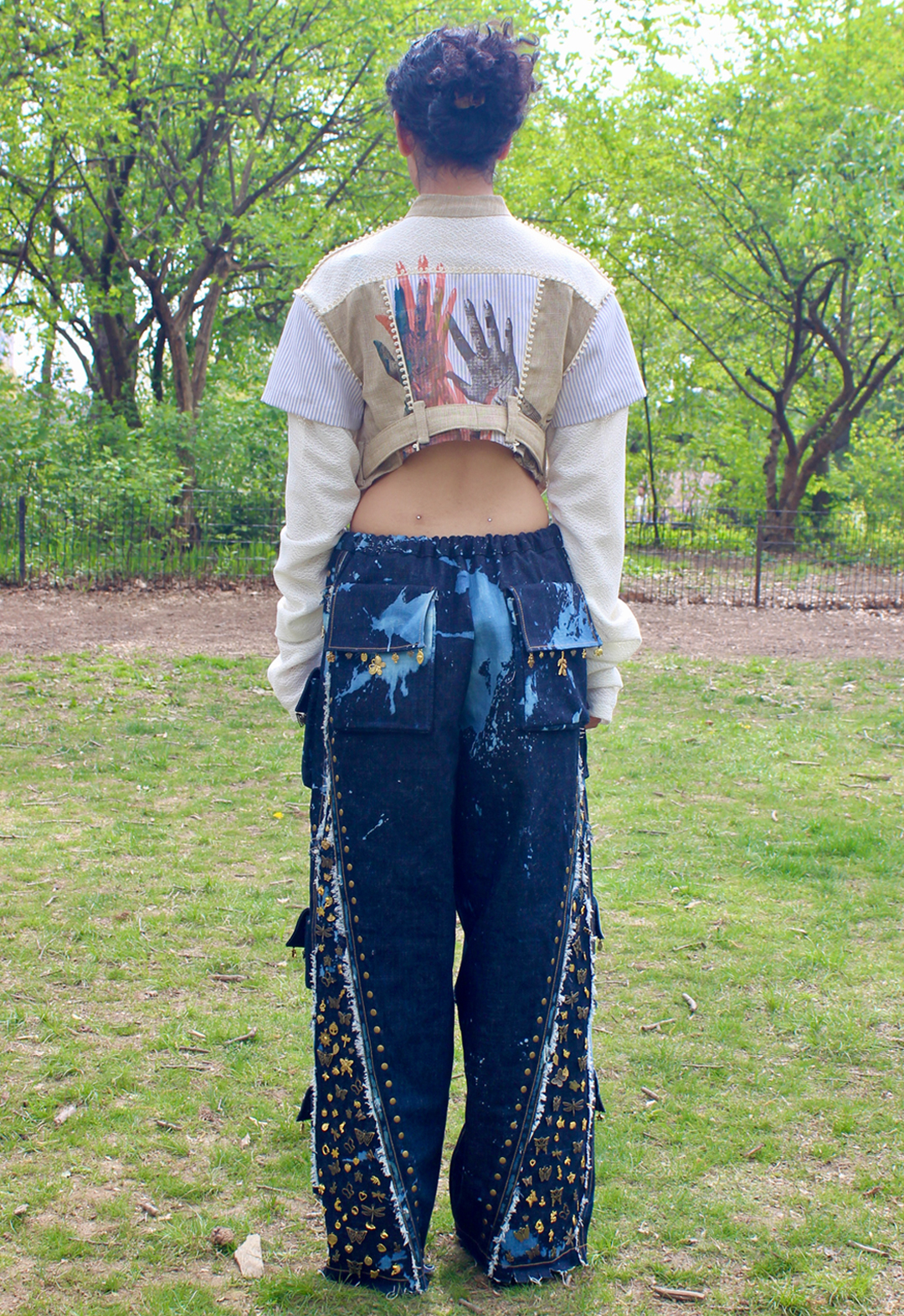 Back view of cropped princess seam button-up top with nehru collar and short and long sleeve layered together. Mixing of three textiles: blue-and-cream striped cotton, mixed tan hues in linen and open weave white cotton blend. Multiple layered screen prints of hands decorated with henna placed in center. Hands are in multiple colors of orange, purple, and blue. Matching tan linen belt. Cream-colored piping with loop shape. Baggy style full-length dark-blue jean with abstract bleaching and cargo pockets. Diagonal style lines with flat-fell seams and distressed edges. Bronze studding following along style lines. Hand-sewn gold and bronze charm embellishments along pocket flaps and filling side area separated by style line. Charms include many shapes, such as butterflies, other insects, and flowers. 