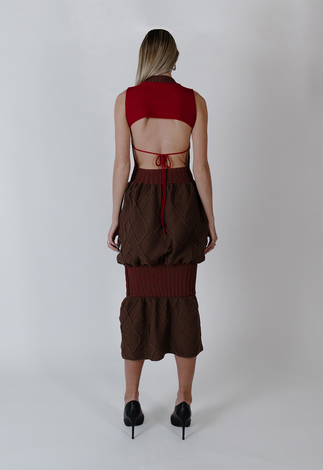 This is a photo of the back of the dress, with an open-back design. The top of the dress is open back, showing the lower back. There is a bow detail in the middle of the model's back. The bottom of the skirt has the same details as the front. Irregular Aran stitches fill the skirt with a balloon shape, creating an exaggeration of the hips. 