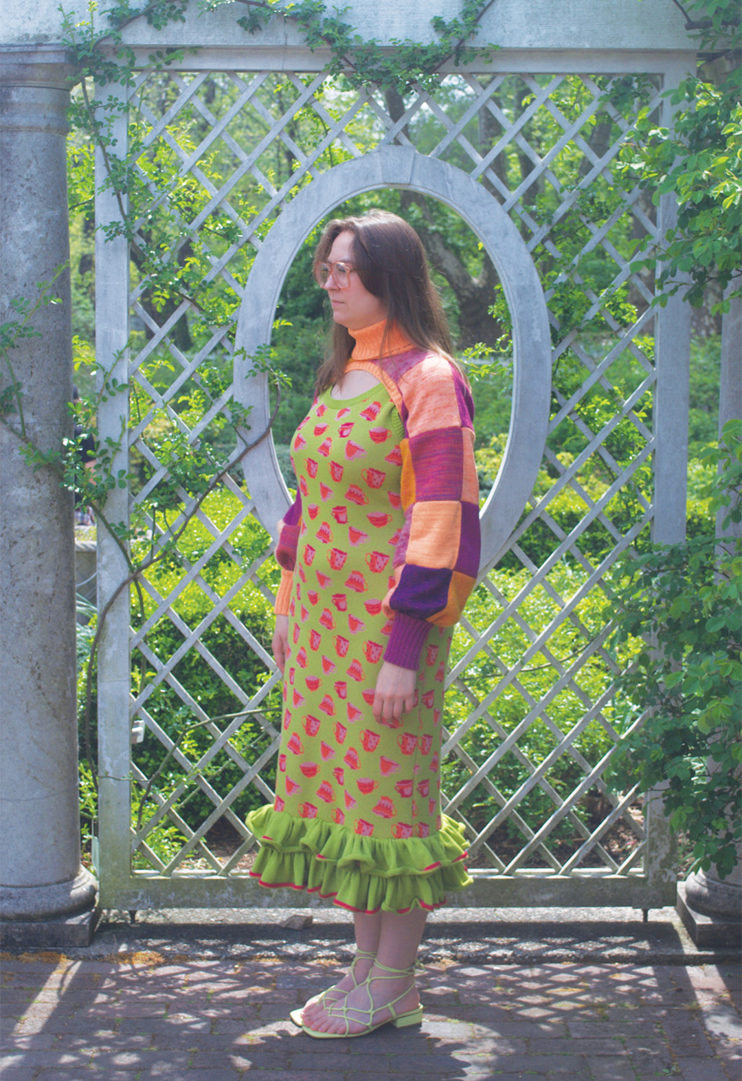 Three-quarter view photo of a model wearing a green teacup motif dress with a ruffle hem and a purple-and-orange patchwork turtleneck shrug. She is standing in front of an oval-shaped window.