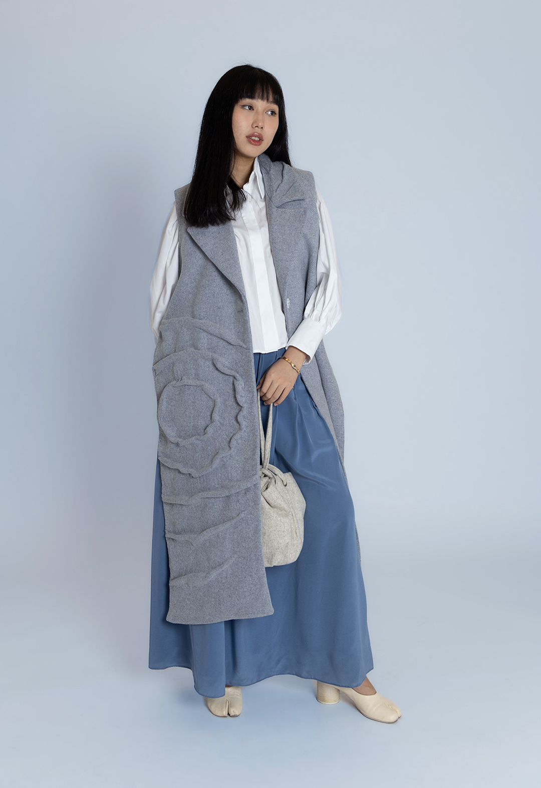 A model presents the front view of a long vest with a ripple texture, layered over a white cotton shirt and blue silk skirt, accompanied by a light-gray pouch bag.