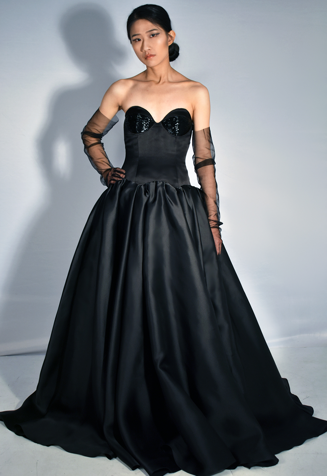 This black gown is made from 100 percent silk. The photo shows a front view of a woman wearing a black silk gown with beading covering three-quarters of the bodice cups. The floor-length gown has a drop-waist duchesse satin bodice with cone-like satin-faced organza skirt panels gathered along the drop-waist, pooling onto the floor. The model is wearing black tulle gloves that cover three-quarters of her arms. Her face is facing the viewer, and she has one hand on her hip. The background is white.