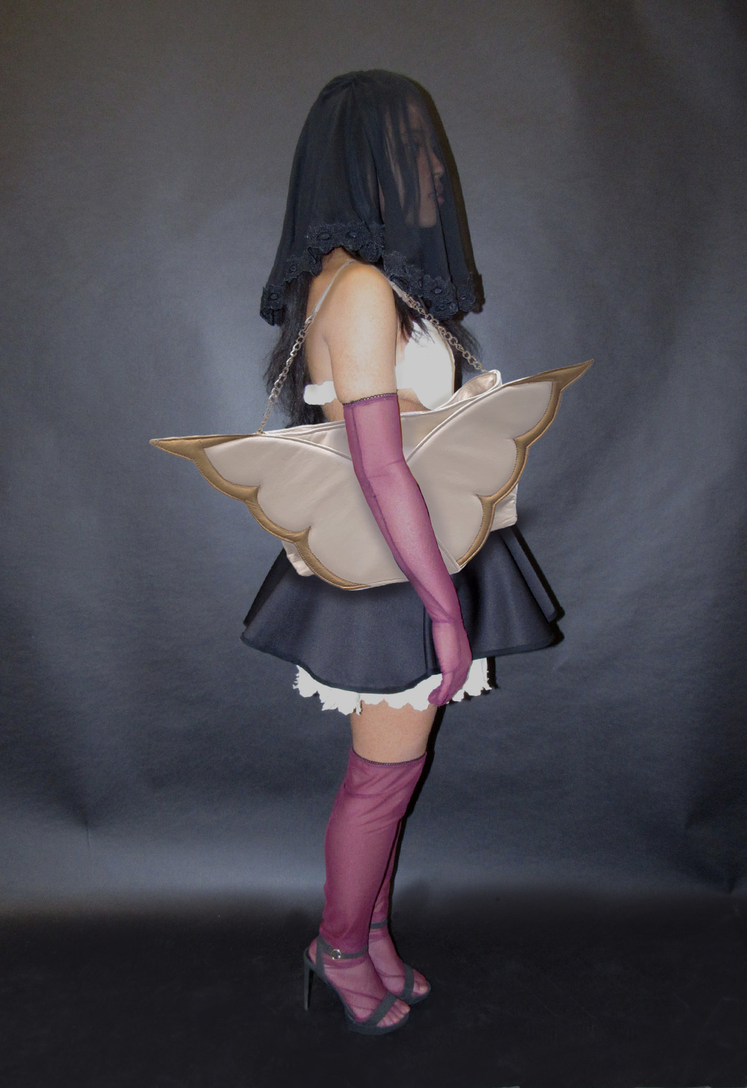 Side-view, full-body shot of a model. She wears a black draped, embroidered veil, an ivory heart-shaped bustier, a black-and-white two-tiered miniskirt, deep-pink gloves, and thigh-high socks. She is holding an oversized ivory-and-gold tote bag with angel-wing motifs.