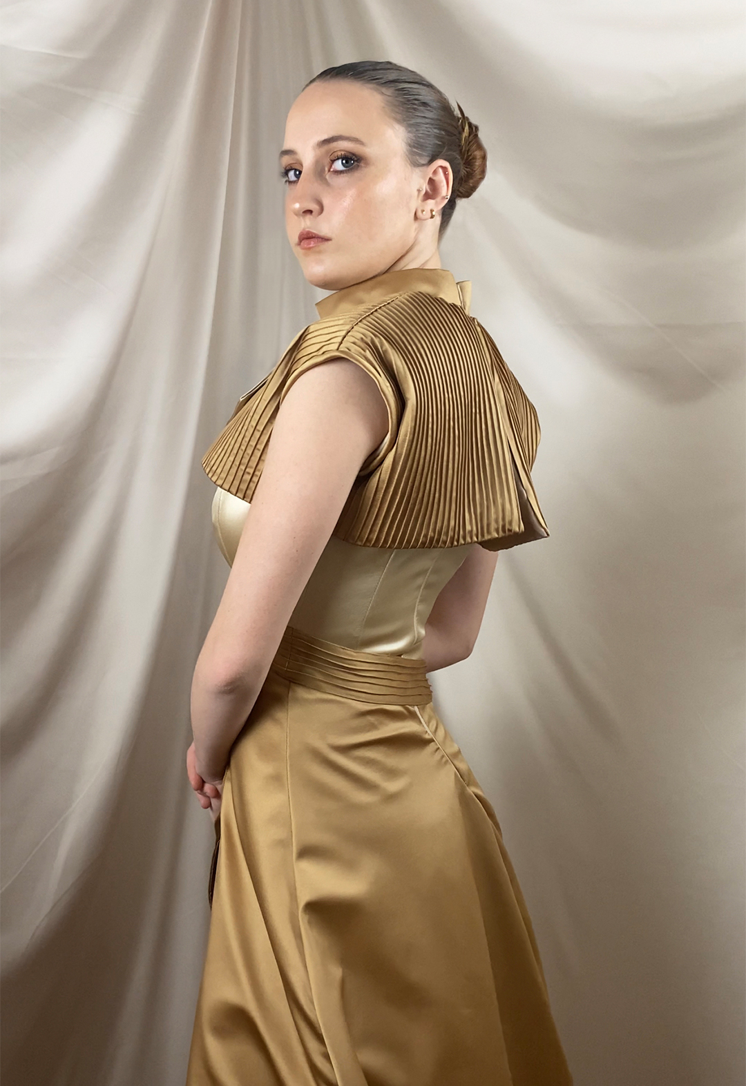 A model is standing in a three-quarter side view, showing the back of the garment. The pin-tucked vest is in full view, and the asymmetrical back can be seen.