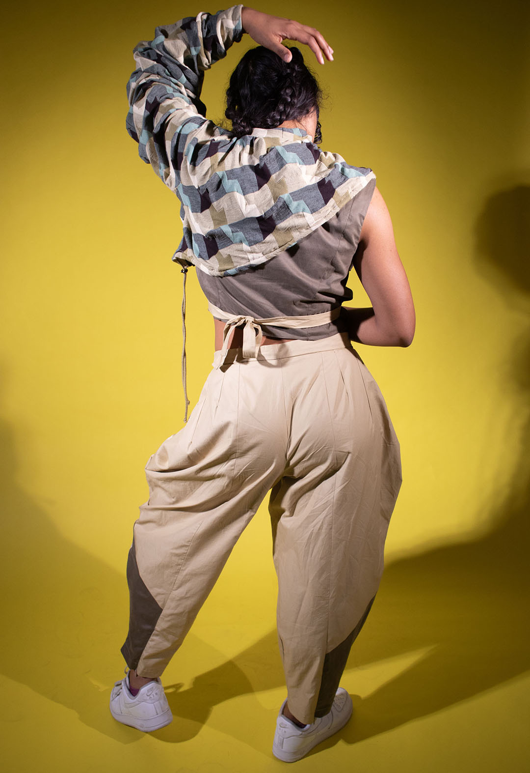 The back view of a young woman in a studio, striking a pose with one arm bent above her head. She is dressed in khaki-colored pants and a matching top, complemented by a distinctive turquoise color-blocked, one-sleeved shawl that adds a creative flair to her outfit. 