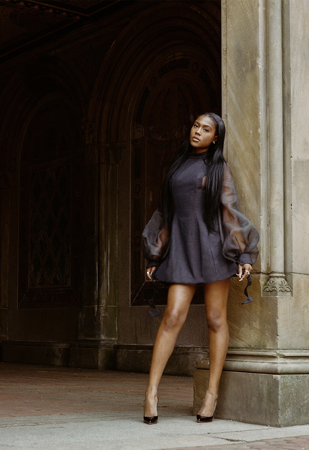 A model standing against a pillar. She is wearing a navy dress with see-through bell sleeves. 