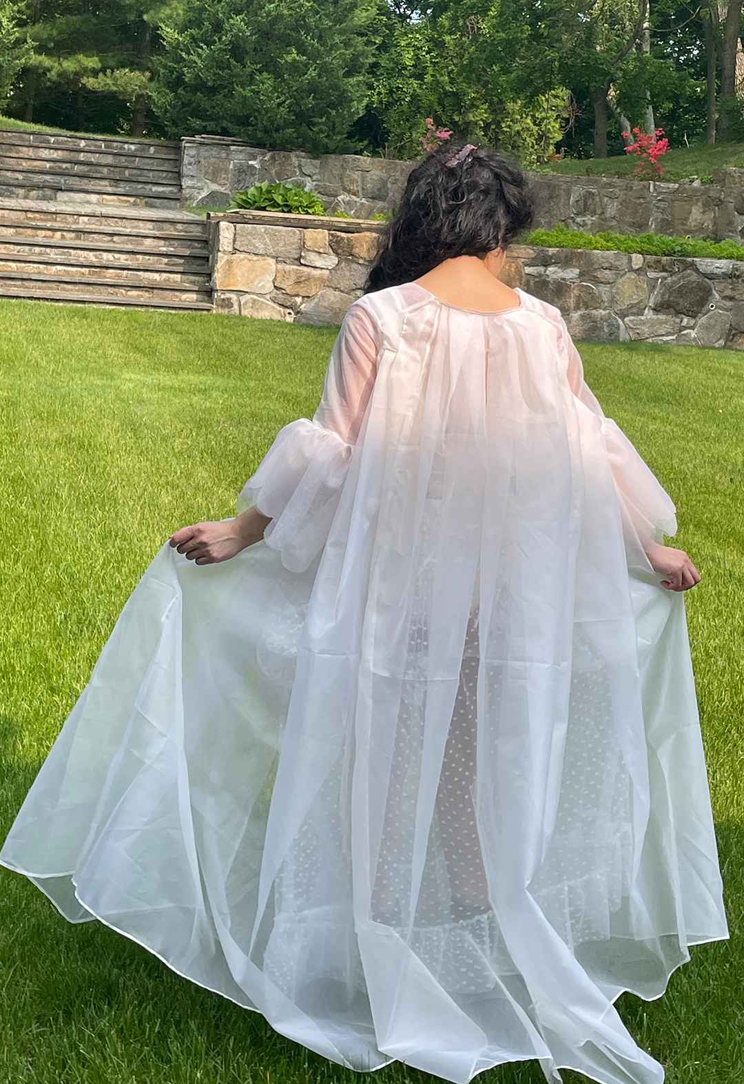 Back view of a model standing in a garden and wearing a translucent off white silk robe with pleats coming from the back neckline.