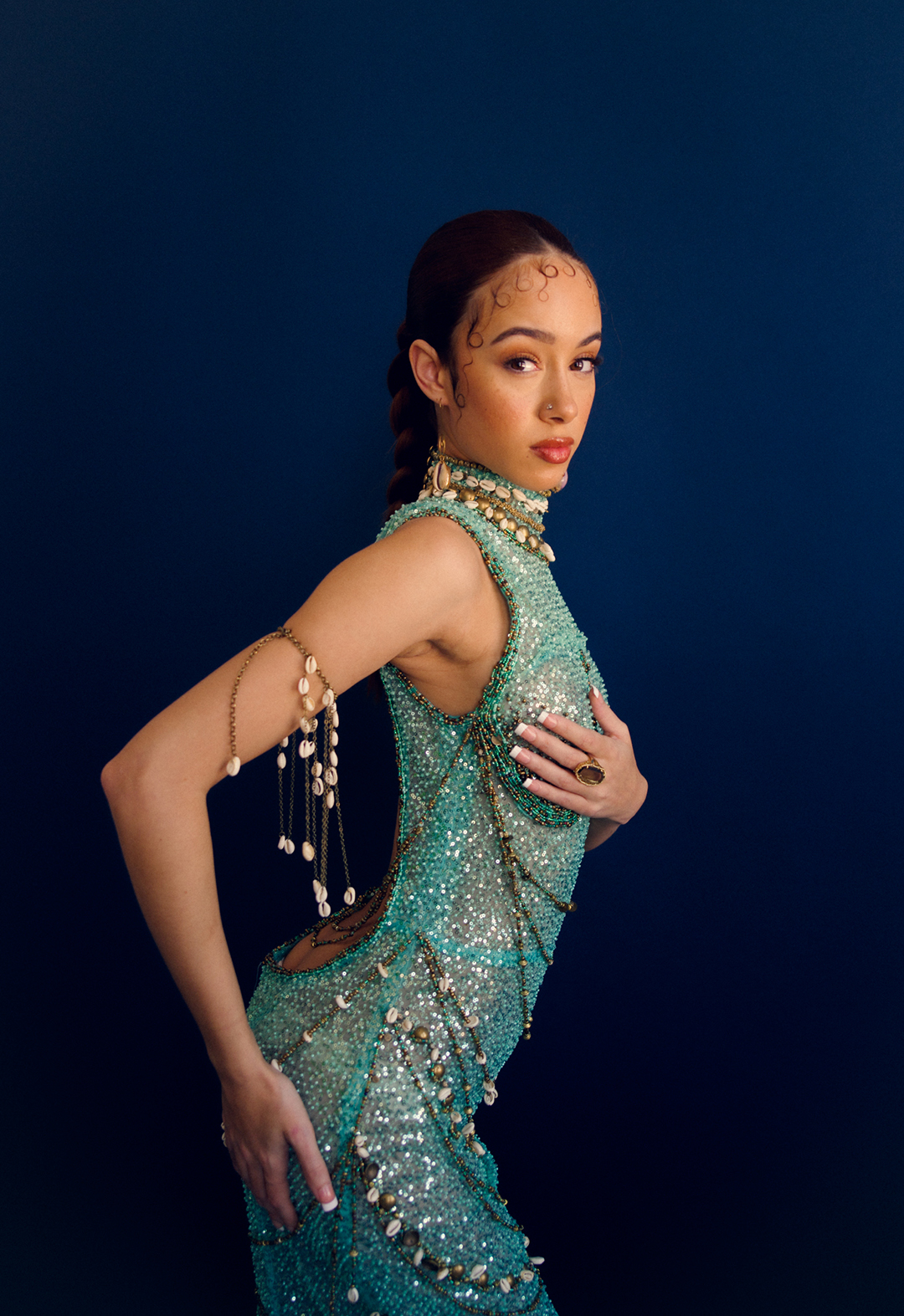 Detailed side view of a model wearing a turquoise beaded dress. One of the model's hands is on her breast and the other one is on her hip. The background is navy blue.