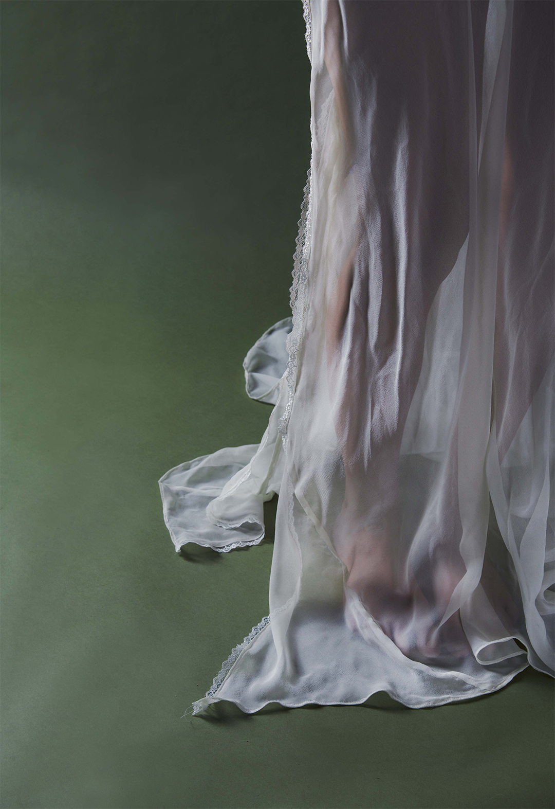 This image features a detail shot of the hem of the pearl-white long bias gown, featuring lace detailing and a rolled hem. The image is taken in a green background setting. 