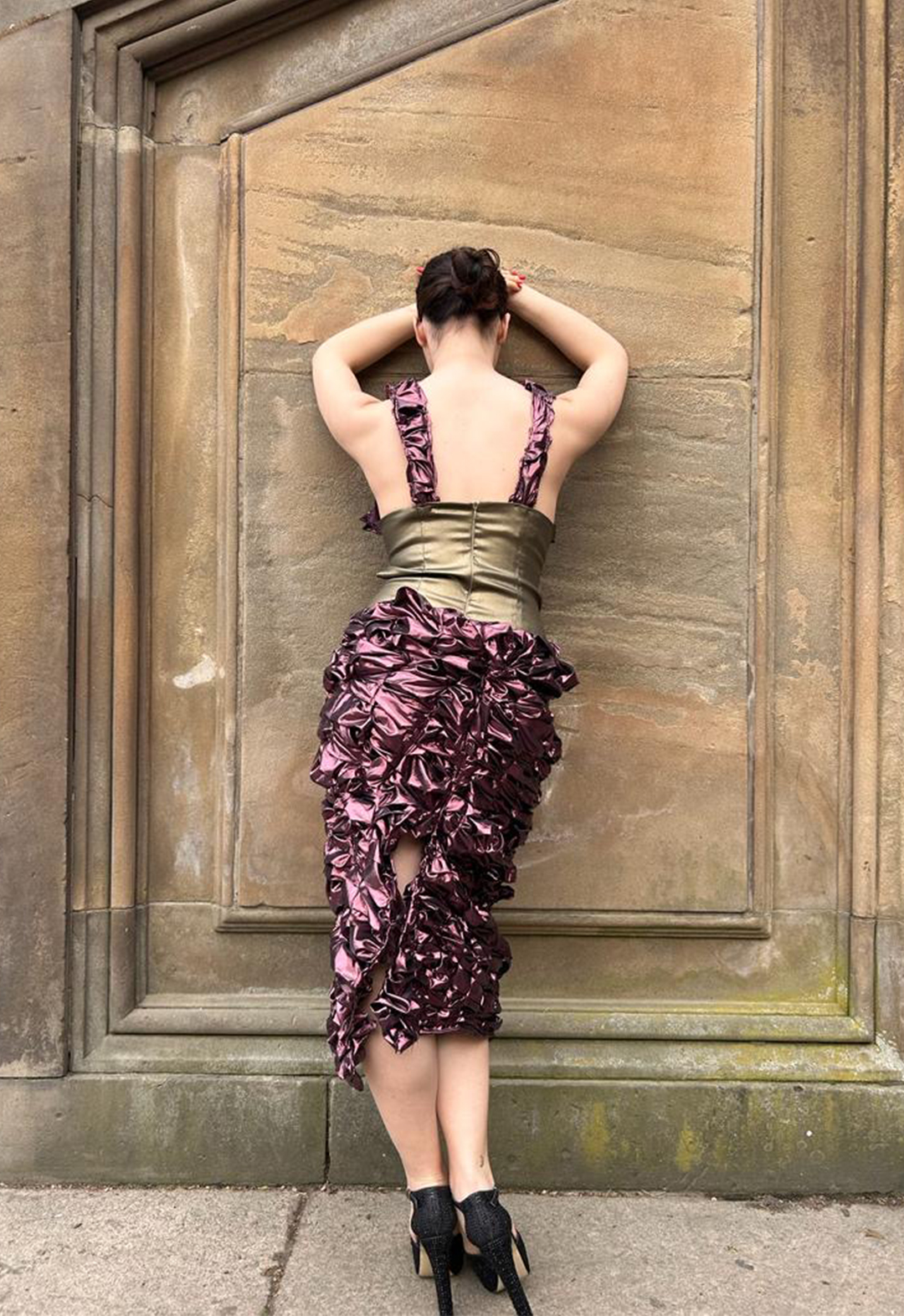 A back view of a girl wearing a long dress and facing an ancient concrete wall. Her hands rest against the wall and come between the wall and her face.