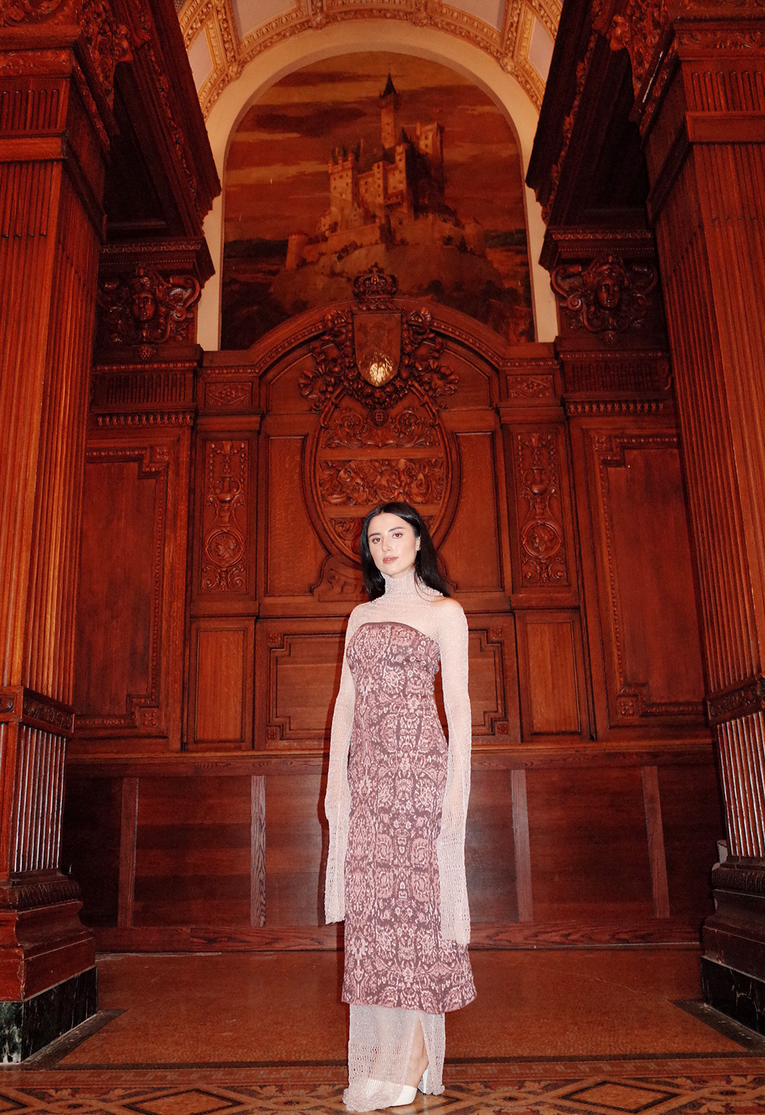 A model standing in a room, wearing a carpet inspired tube dress layered over sheer pointelle dress. The walls in the background are covered with wooden carved columns.