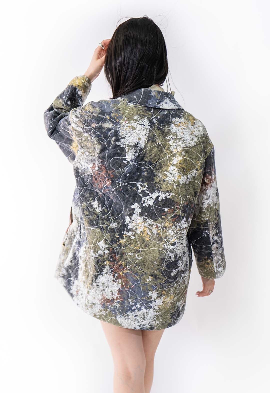 This is a back view of a jacket that is made from chopped-up scrap fabric, all stitched together with white thread and a small zigzag stitch to create a colorful abstract pattern of whites, blues, greens, and burnt orange. The jacket has a straight boxy shape. The model is standing in front of a white background. Her hair is pulled forward to one side, and she has one hand running through it while her other hand falls straight by her side.