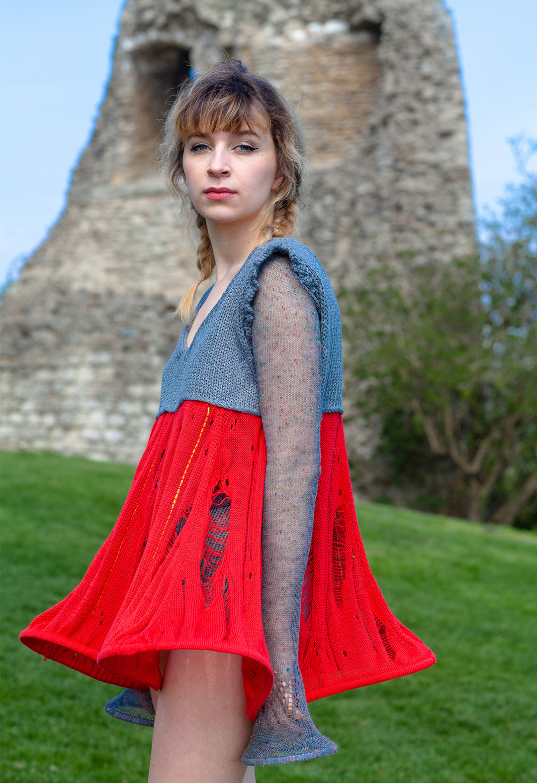 A close-up image of the model looking toward the camera but standing sideways. She is wearing a gray-and-red knitted fairy-tale dress, gray knitted briefs with a crochet edge, and a sheer lace top with bead accents.