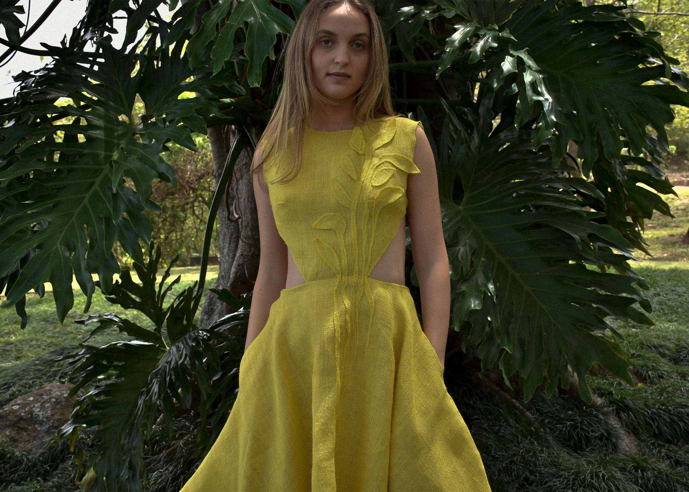 Yellow burlap dress, hand-embroidered with yellow burlap leaf appliqués. Model is standing in front of a big tree full of monstera leaves.