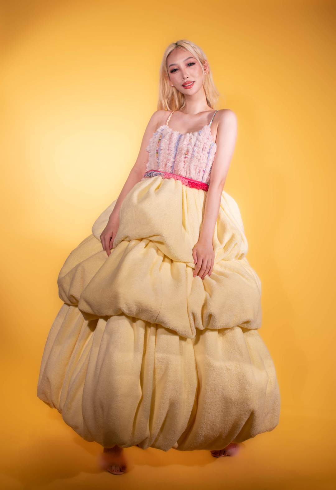 Photo of a girl with blonde hair wearing a pastel pink-and-yellow bubble dress. She is smiling and both of her hands are on the dress. The dress has two stripes with different colors, a light-pink furry bodice, a bright-pink knit belt and a light-yellow three-layered bubble skirt.
