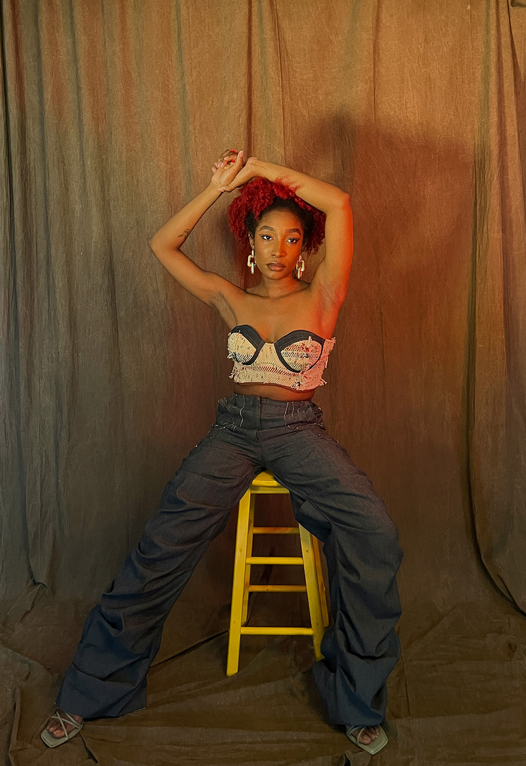 A model is sitting on a stool with her arms above her head in a woven bustier and denim pants.