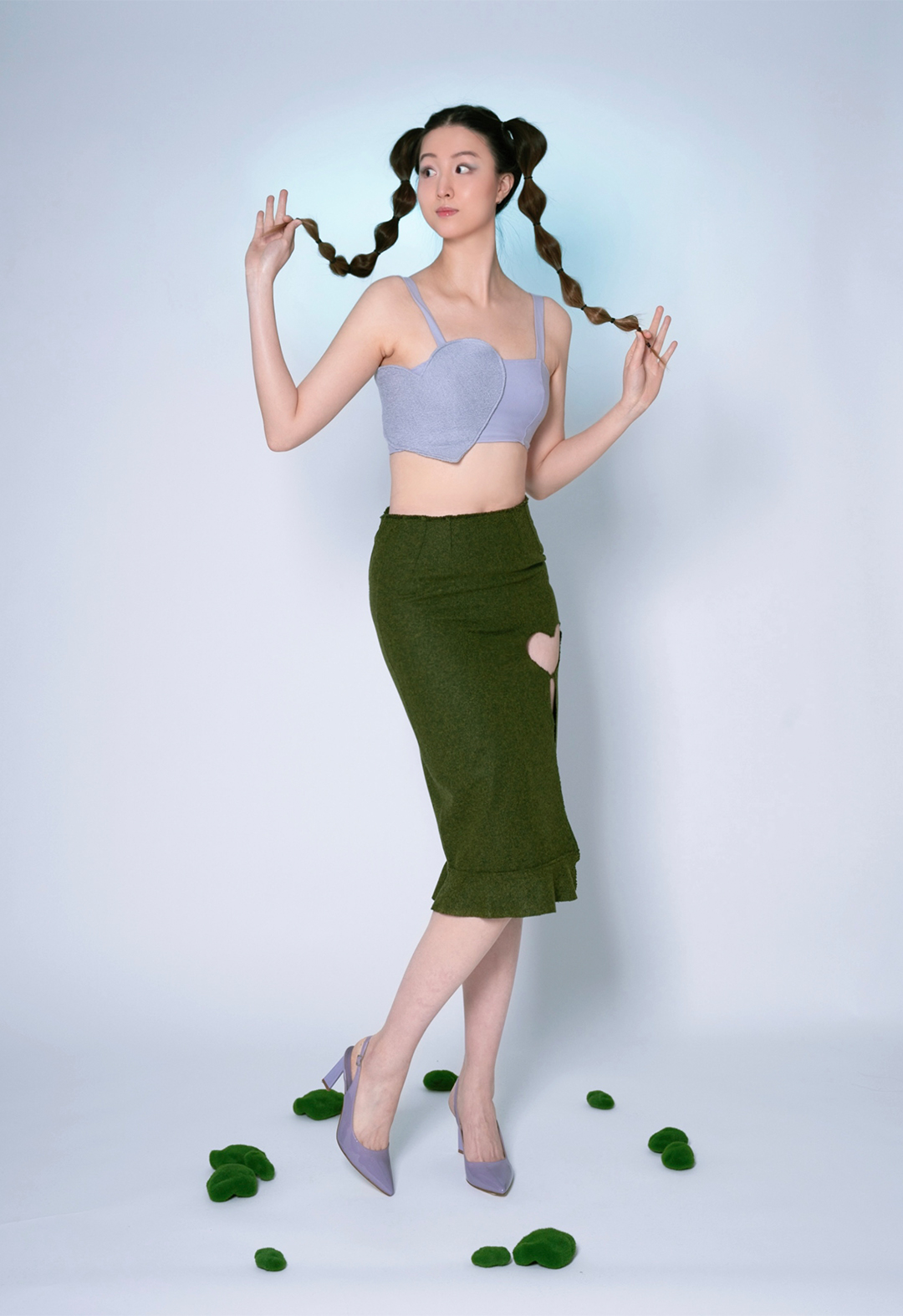 A three-quarter view of a model playing with her hair, which is styled in two bubble ponytails. There is a purple heart patch on the right side of the top, a heart-shaped cutout and split on the right side of the moss-colored skirt, with a ruffle at the hem. 