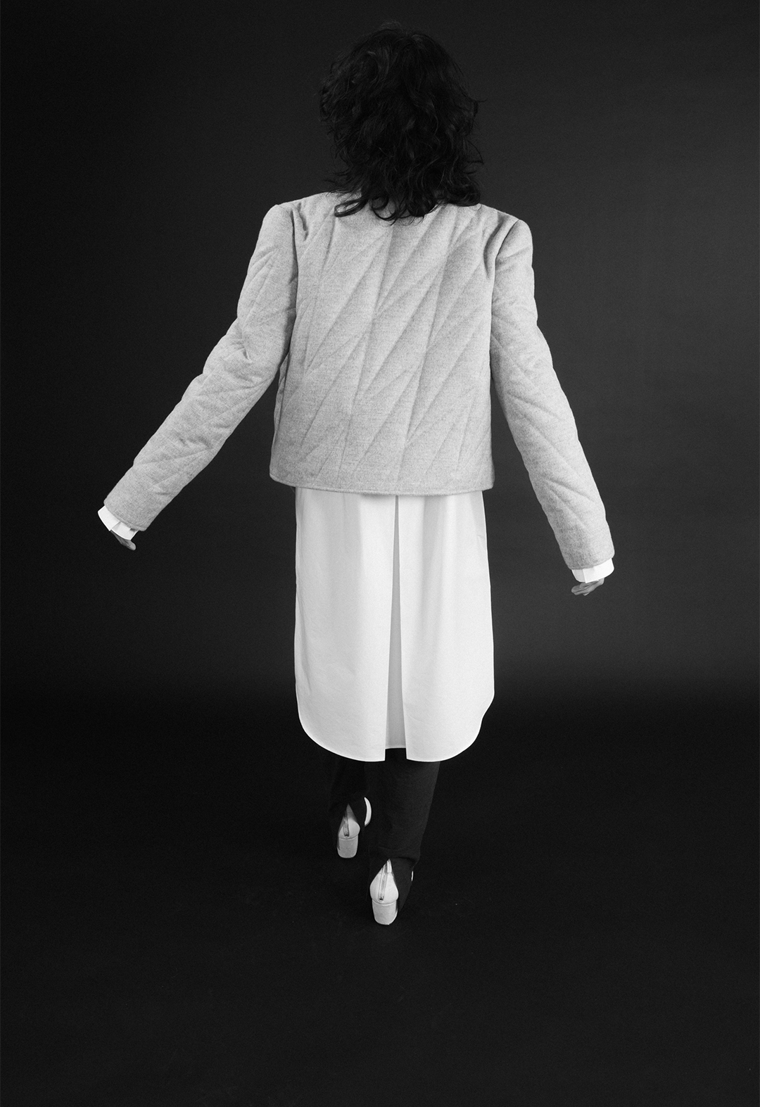 Photo of the model, Aria Puga, wearing a grey wool quilted jacket, a white cotton tunic shirt, and teal linen trousers. The jacket has geometric quilting. The back of the tunic shirt has an inverted box pleat. The trousers have a back slit at the hem. The model is turned with their back to the camera. The background is black. The photo is monochrome.