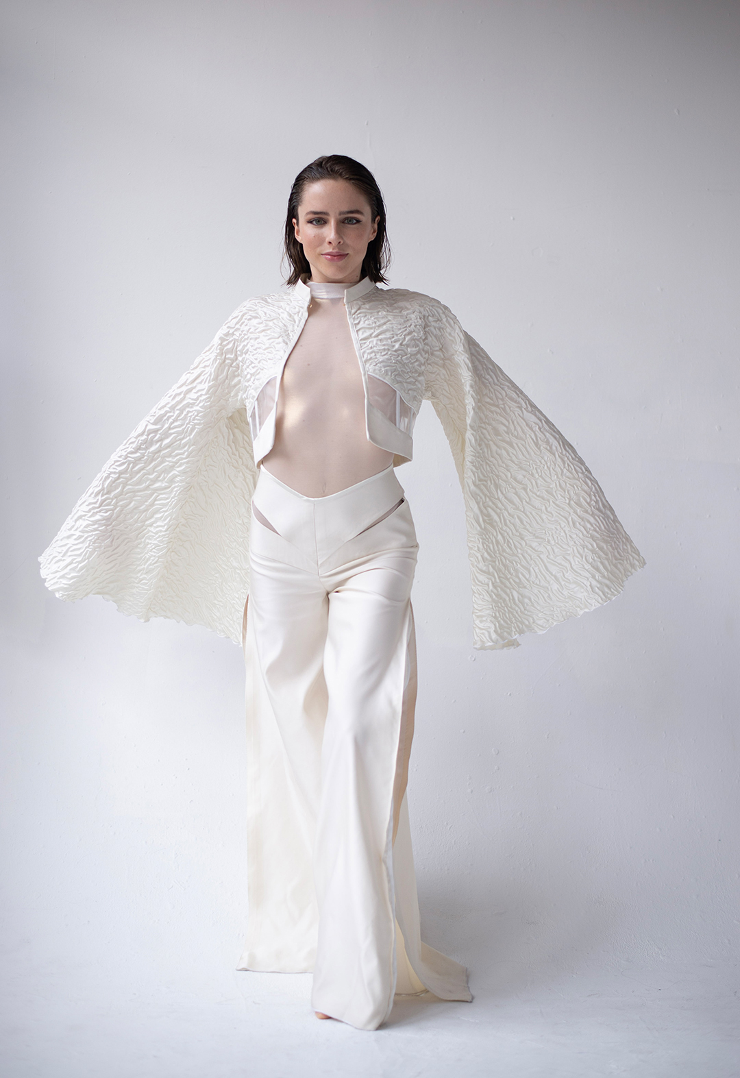 Front view of a model wearing a seashell white corset jacket with flowy kimono sleeves and flared, cut-out, and slitted pants. Her arms are stretched to her side like wings. 
