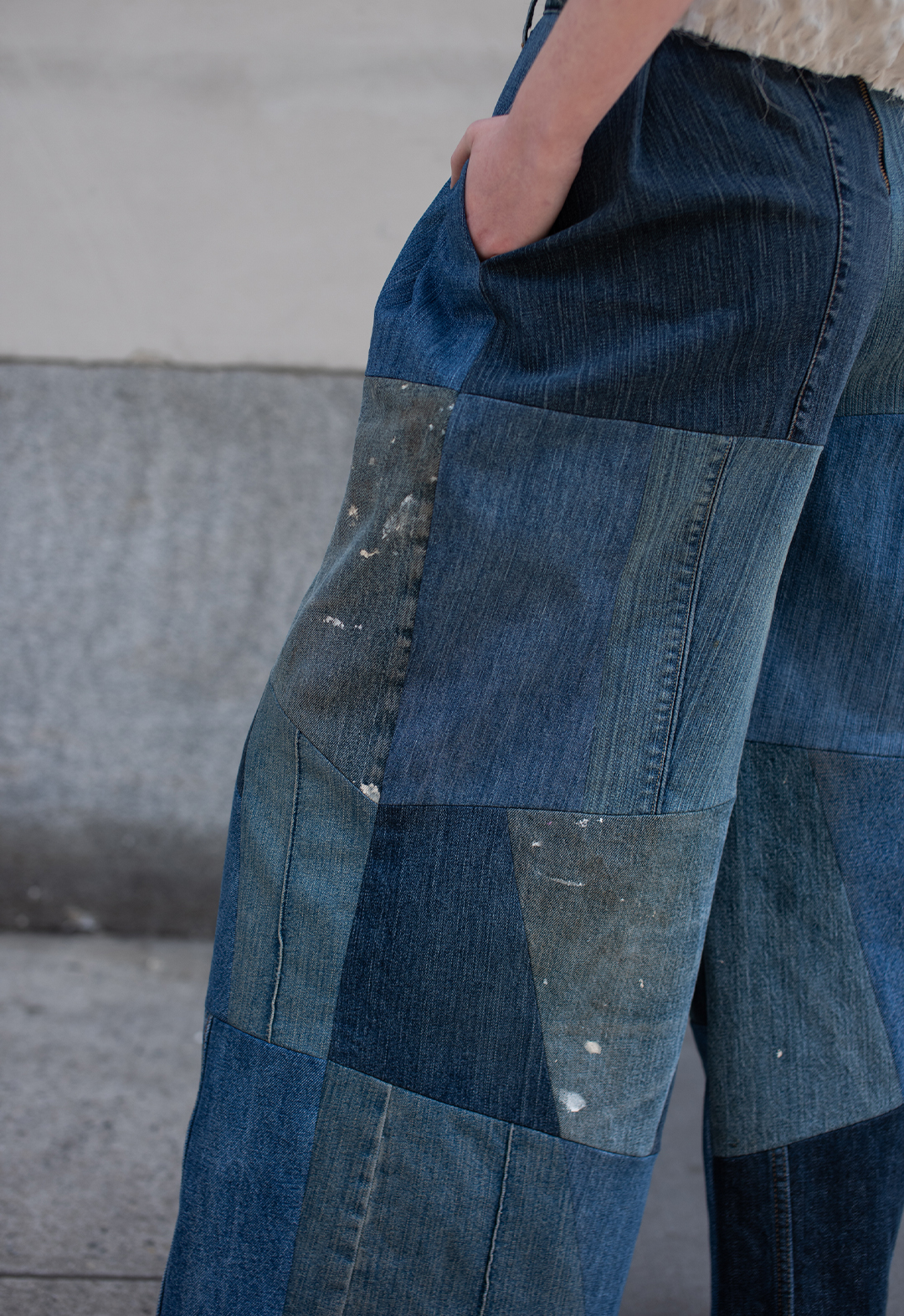 These pants are made by upcycling denim jeans and repurposing the fabric into a patchwork layout, as well as utilizing the inseam of the jean to create a design detail that enhances the assortment of denim-blue washes. The photo is of a girl's bottom torso with her hands inside the inseam pocket, one of her legs extends slightly outward, revealing the patchwork design. The background has multicolored gray and beige concrete panels.  