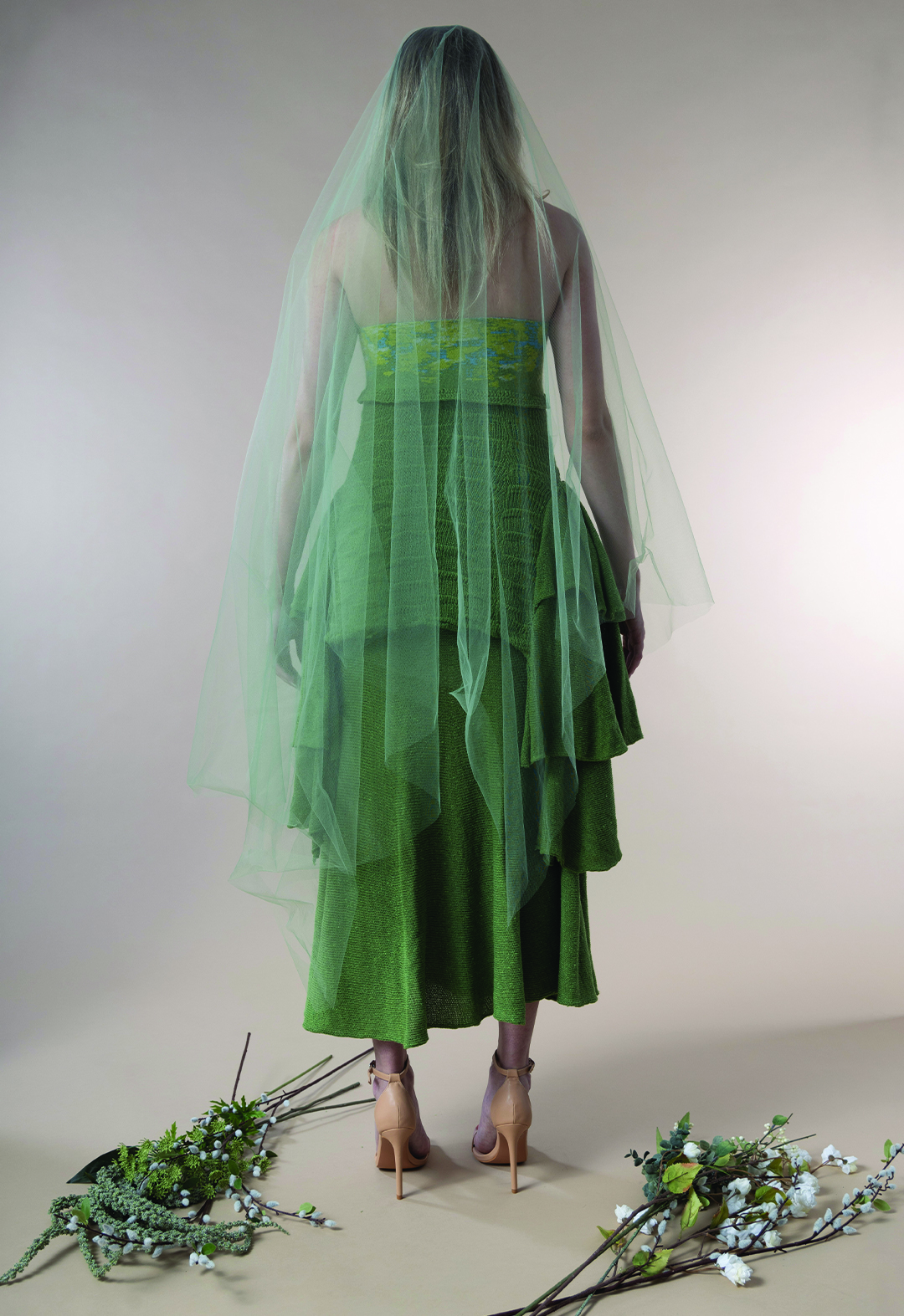 The female model is shown from the back. She is wearing the heather-green knit gown, and a translucent green veil covers her from the top to the knee level. 