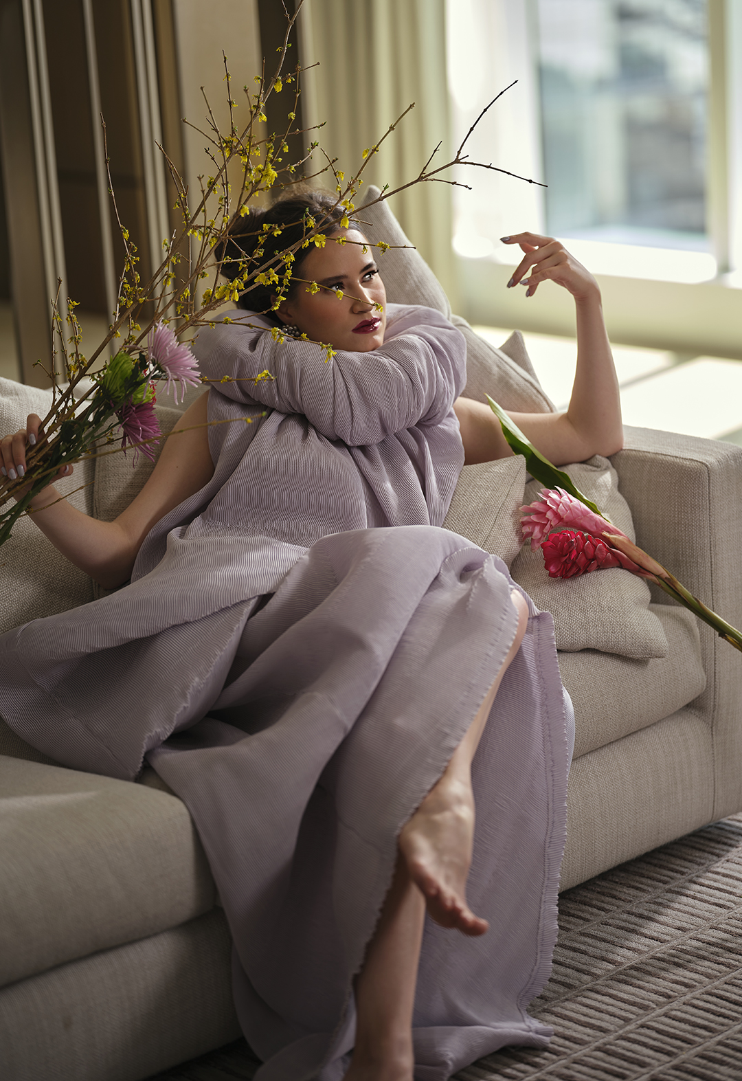 A female model in a purple pleated balloon dress with a train and a separate puffy collar is sitting down on a couch, with her left arm raised at a 90-degree angle and her right arm open to the side. In her right she is holding purple and pink and green flowers and branches with small yellow flowers. There is a big pink flower with a big green leaf lying on the couch on the model’s right side.There are multiple cushions behind her back to help her sit straight. The pleated fabric allows unique folds on the garment when sitting down.