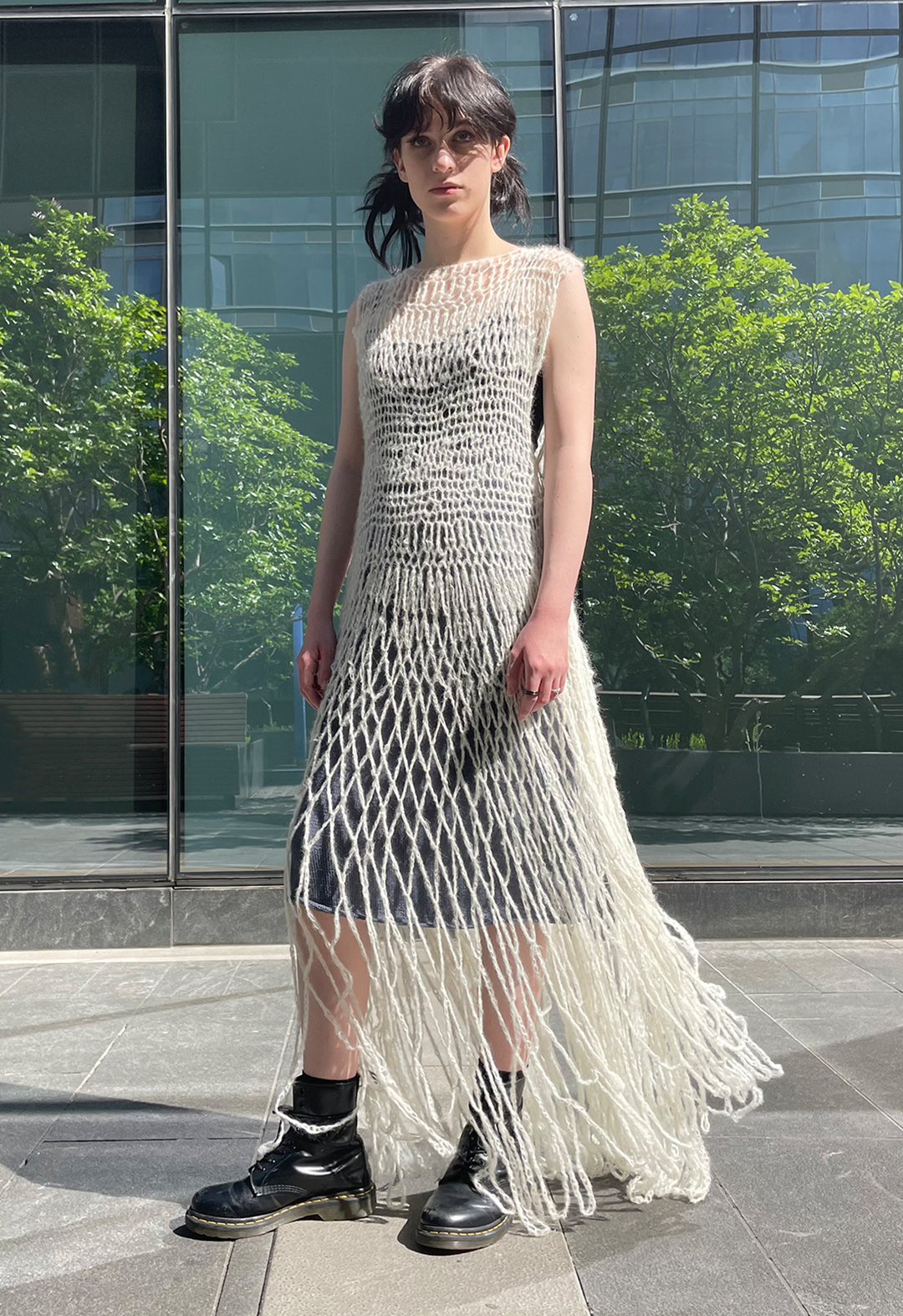 Front view of a model wearing a hand crocheted white lace suri alpaca dress. The model is wearing combat boots and there is a glass building in the background, with reflection of trees.