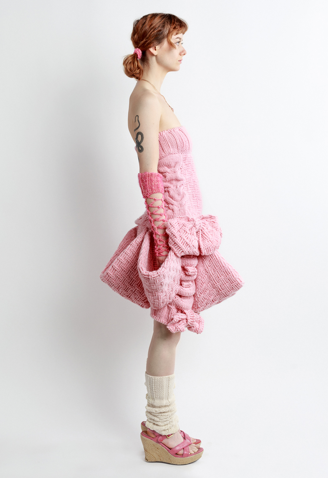 Photo of a girl posing in a pink sleeveless magical girl mini dress, looking to the side. The dress is hand knit with baby pink wool. The dress features big bows and big pockets circling the skirt of the dress. The dress, and girl, are posed at a side view to the camera. The girl is wearing crochet fishnet gloves, heels, leg warmers, pigtails, and a heart necklace. The girl has her hands to the side, in two of the big pockets of the dress. 