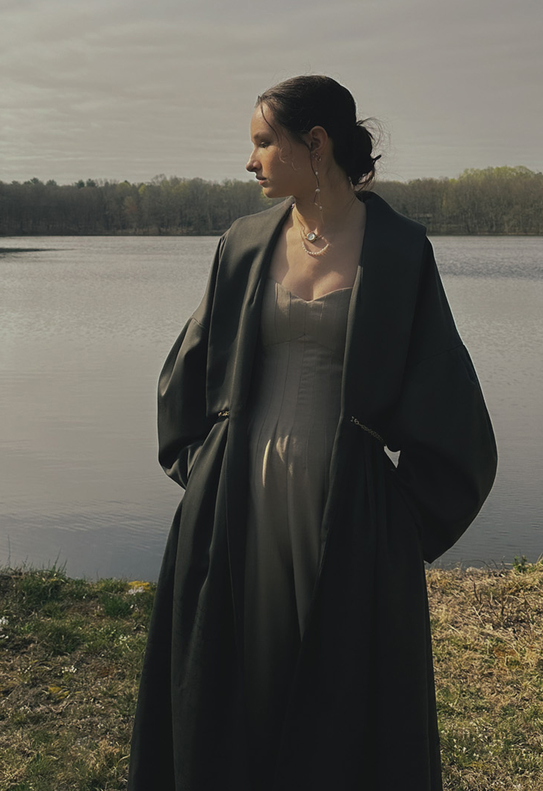 Photo of a girl wearing a long green coat and beige jumpsuit with her hands in her pockets. She is standing in front of a scenic background with a lake, clouded sky, and trees in the distance. 