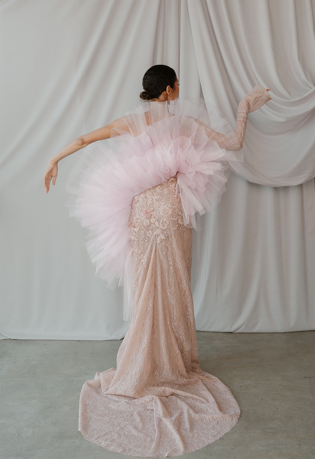 Back view of model in blush lace, one-shoulder sleeve dress with godet train, lace appliqué, and pleated tulle coming from a curved seam over the bust.