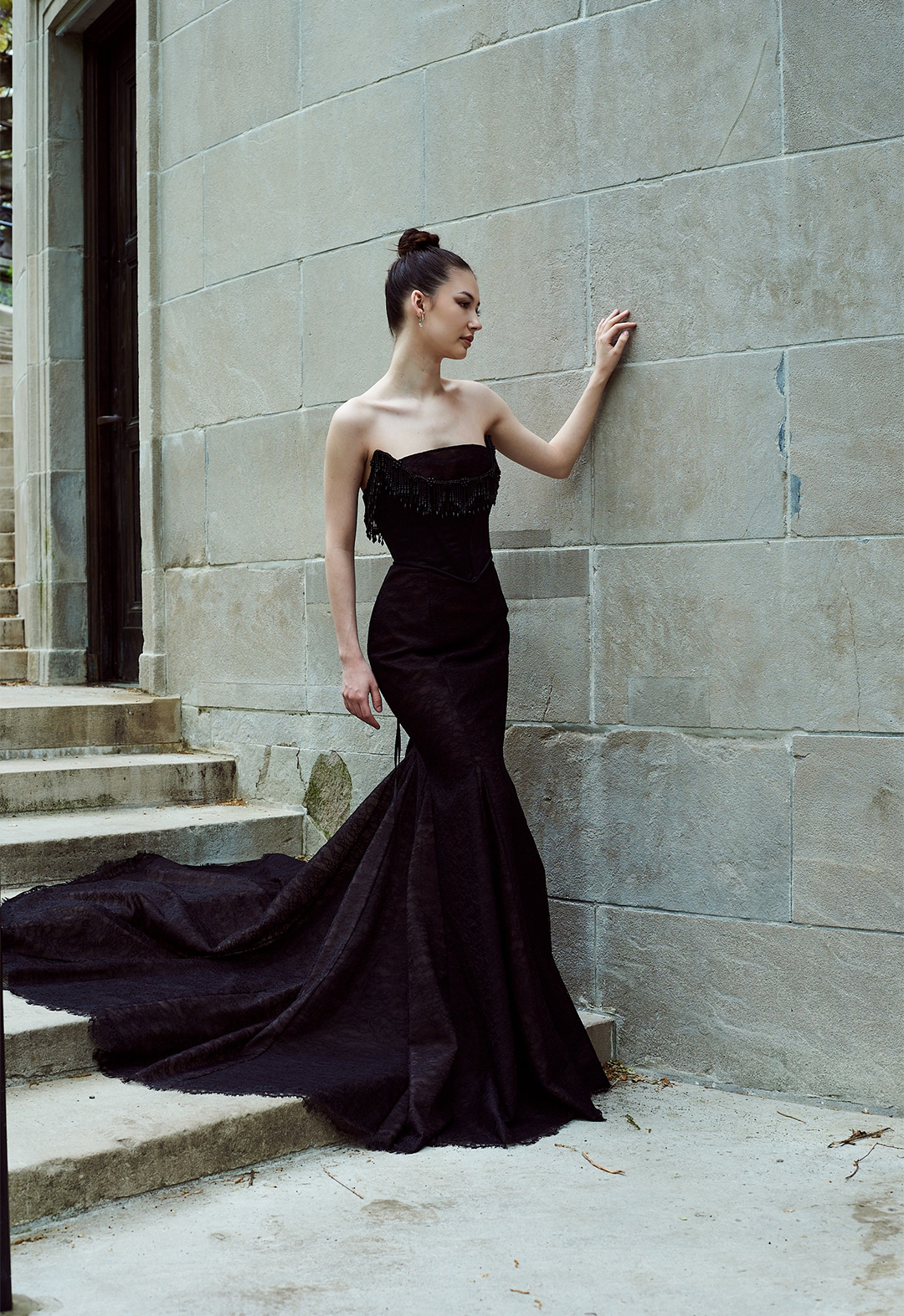 Model walking at a three-quarter angle with one hand on the wall and one hand down. Full body shot with neutral stone background staircase.