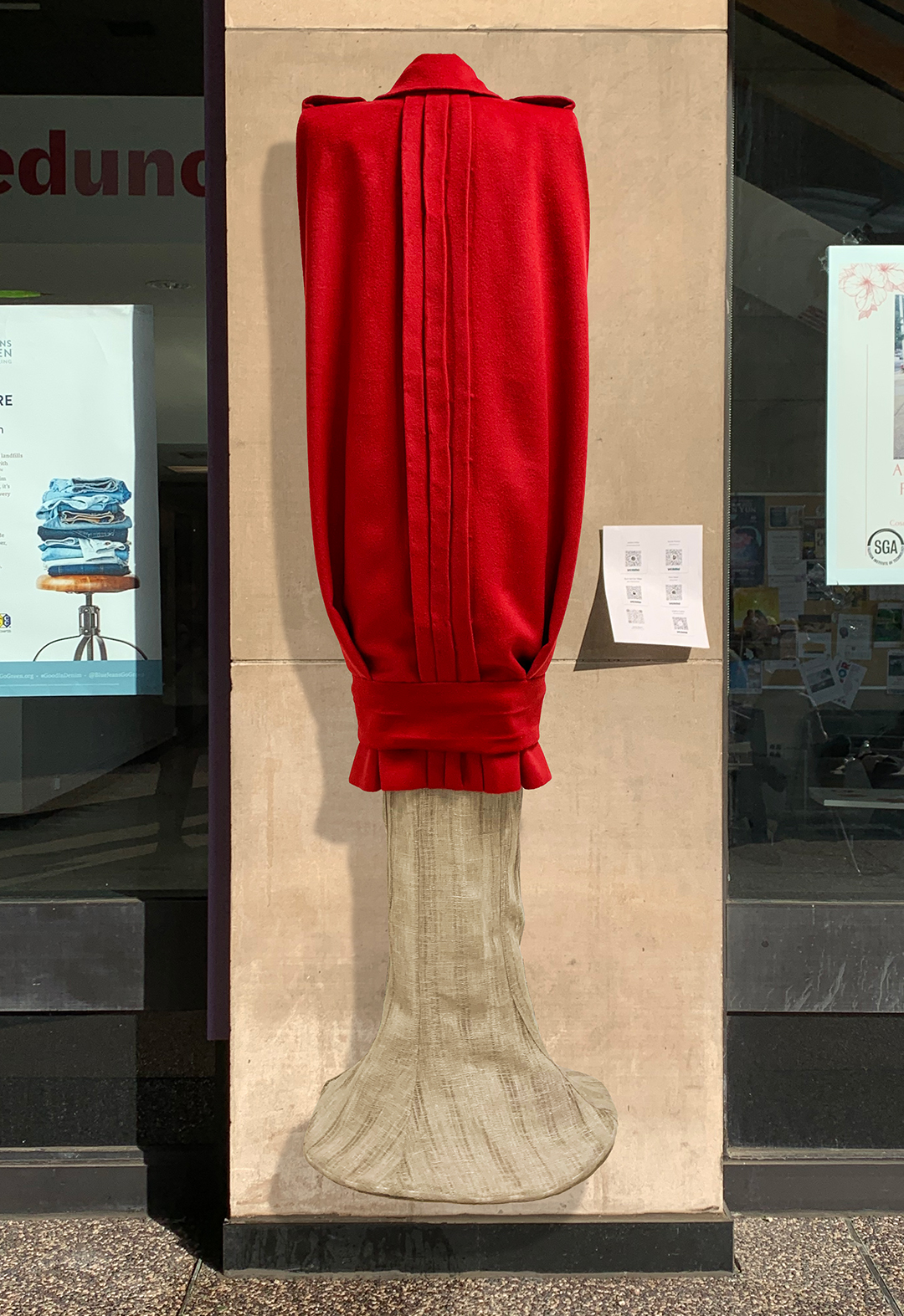 There is the back of a long red wool vest. The linen dress is underneath the vest. The background is concrete column and glass windows. 