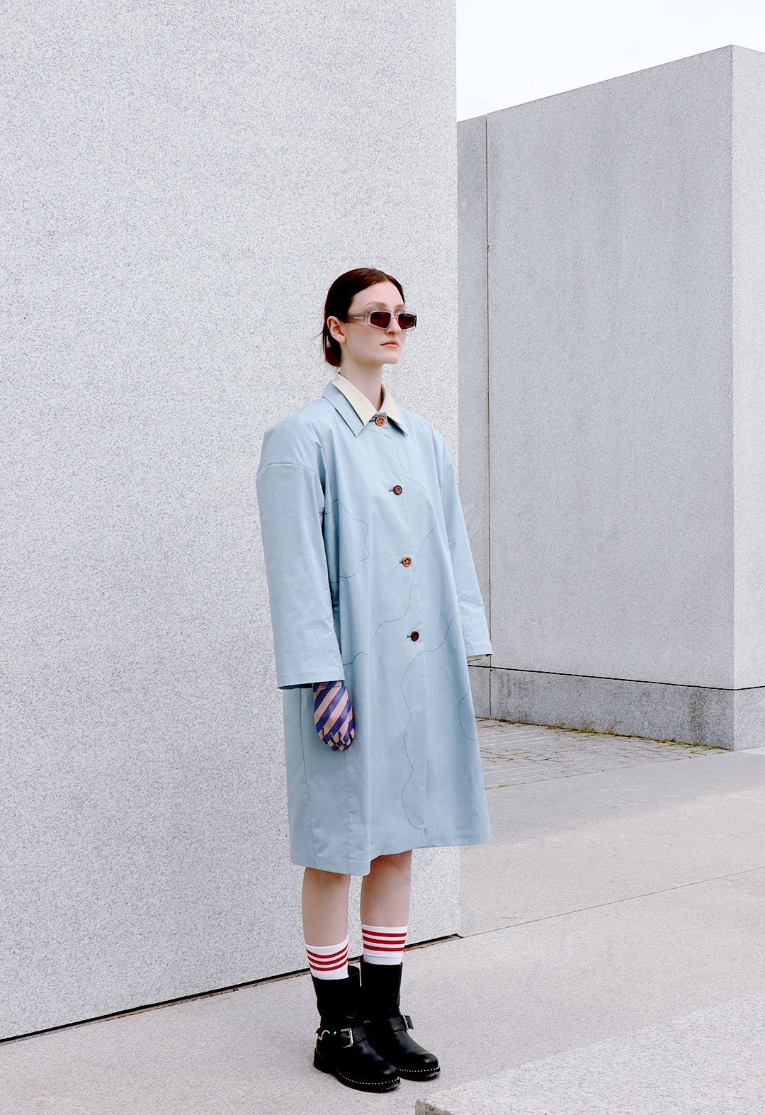 This is a long, blue oversized coat, with flower-inspired embroidery detail and ethnic wooden vintage buttons. It is accessorized with dark-blue mesh long-sleeved gloves, sunglasses, red striped socks, and black boots. The model is posed in front of a gray wall.