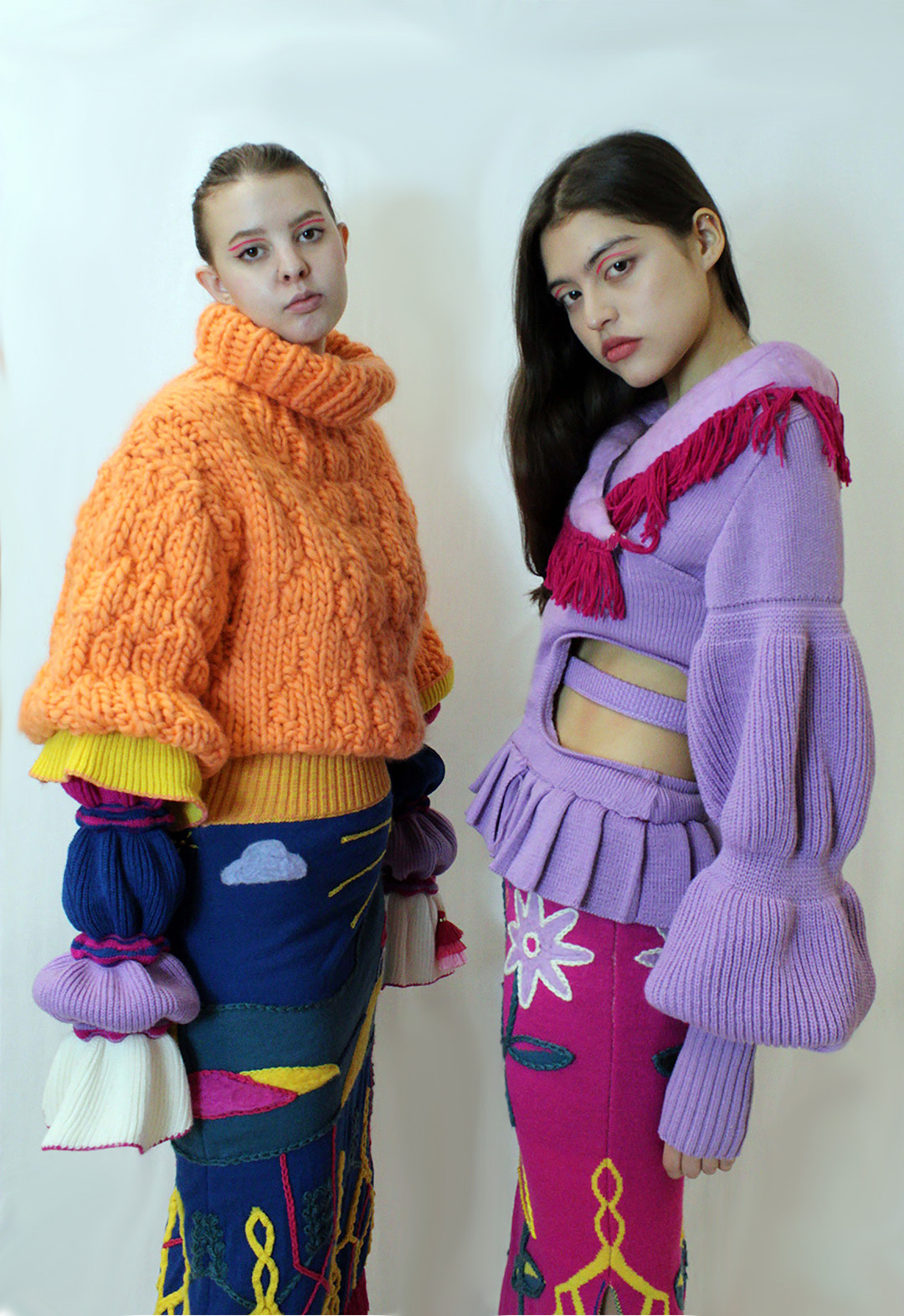 Two models standing in front of a white background. The model with long hair is wearing a lavender-colored knit wrap top with shaped sleeves and crochet fringe at the shawl collar. She also is wearing a skirt with a floral jacquard multicolor pattern. The model with a pony tail is wearing an orange knit sweater with multicolor sleeve. 