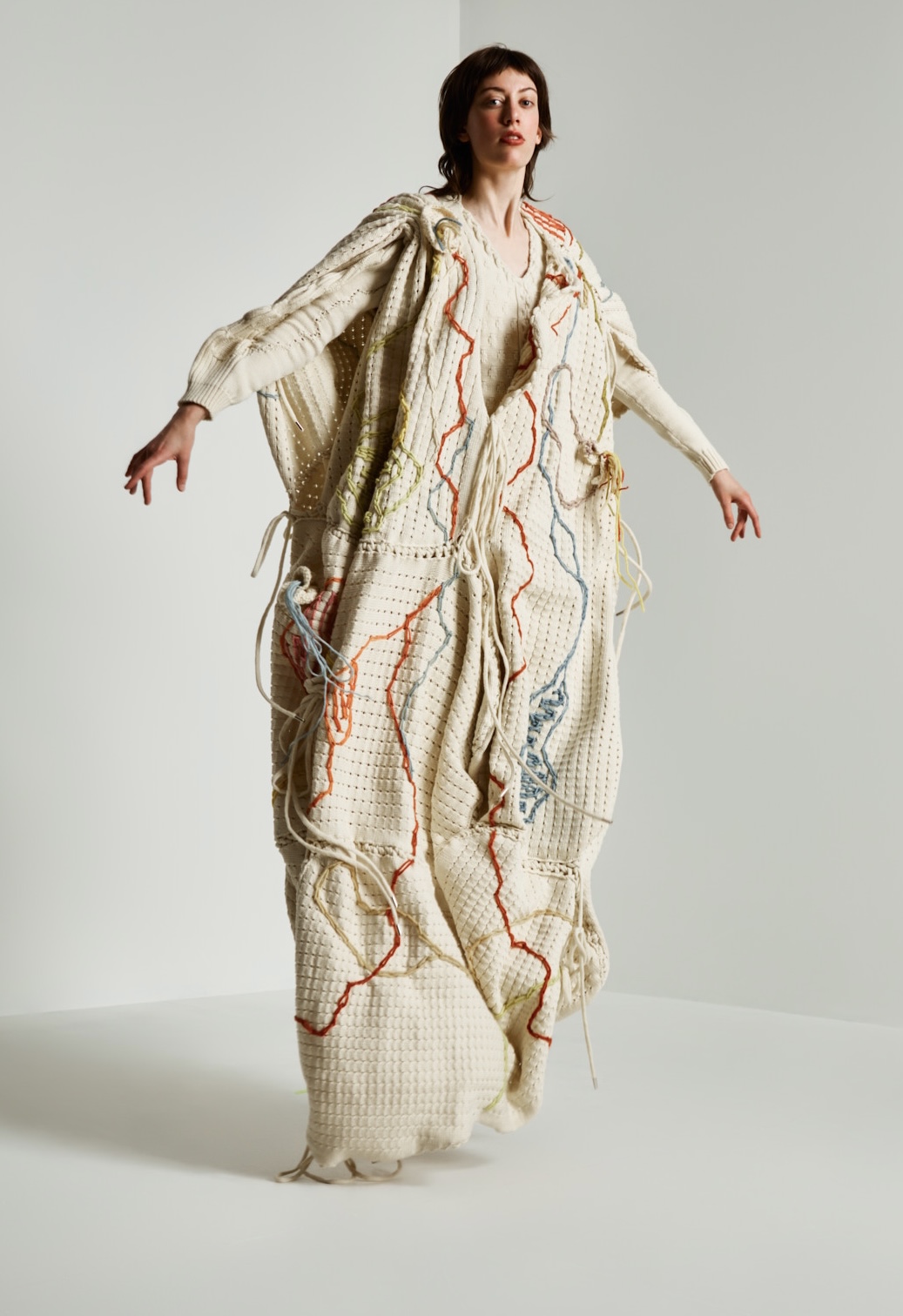 Full-length image of a model posing in a cream knitted garment, featuring embroidery and a handkerchief head covering. She appears to be jumping or floating.