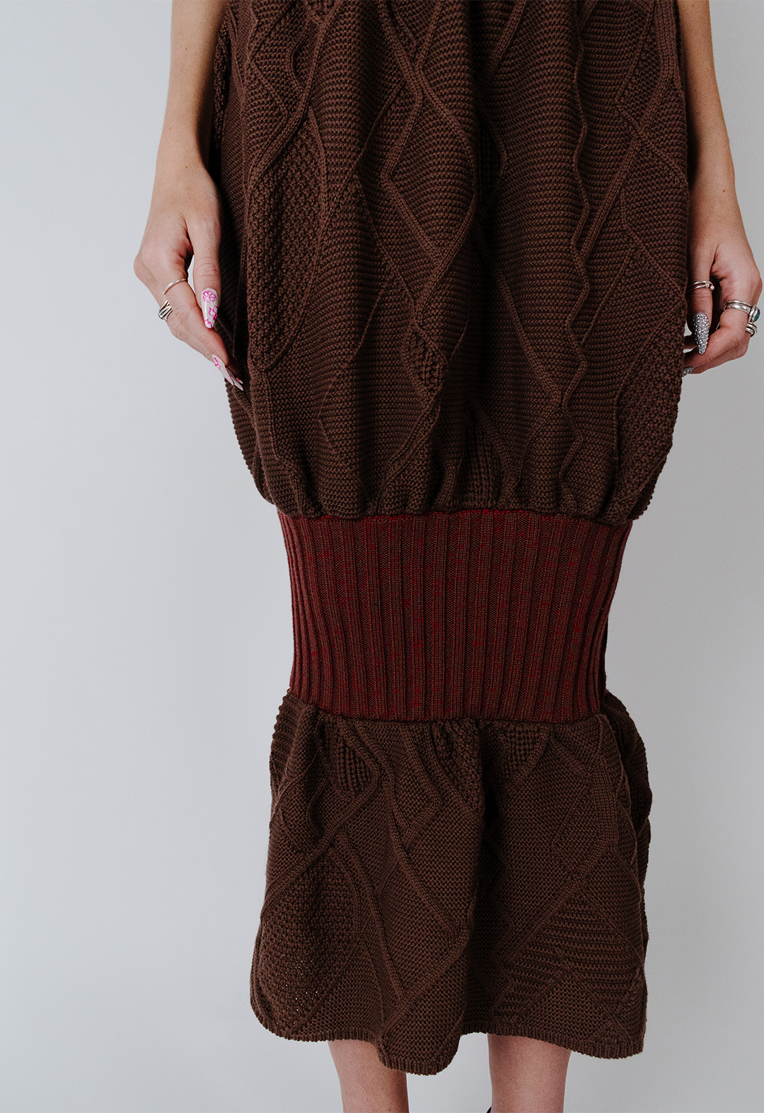 This is a close-up image of the balloon skirt. The model's hands are to her sides. The brown balloon skirt is filled with Aran cables running through it, allowing you to see the mix of colors in the rib. There is a mixture of red and brown on the rib section on the knees. 